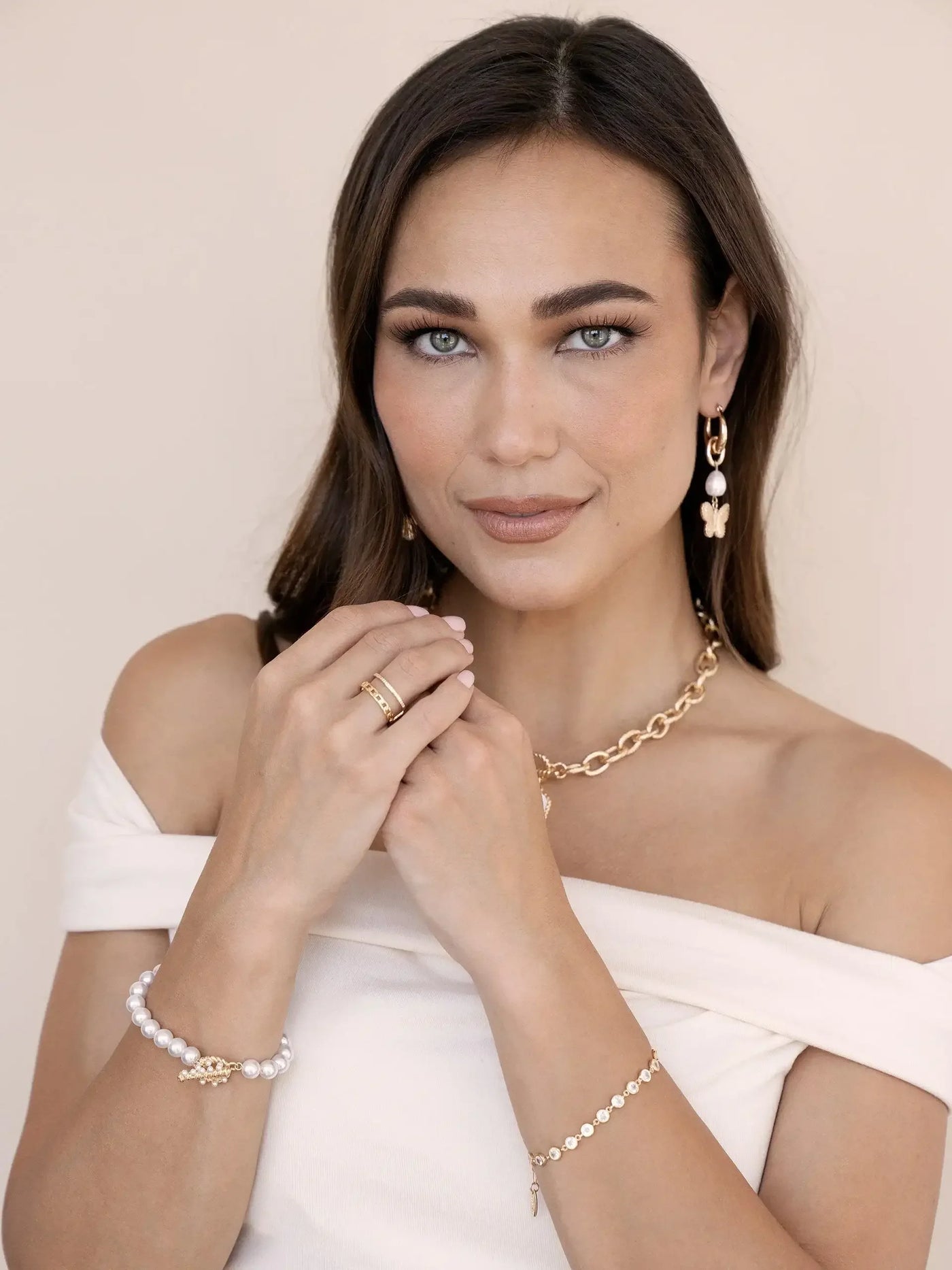 A woman with long, dark hair is gently holding her hands together, showcasing multiple elegant jewelry pieces, including bracelets and earrings. She is wearing an off-shoulder top against a light background.