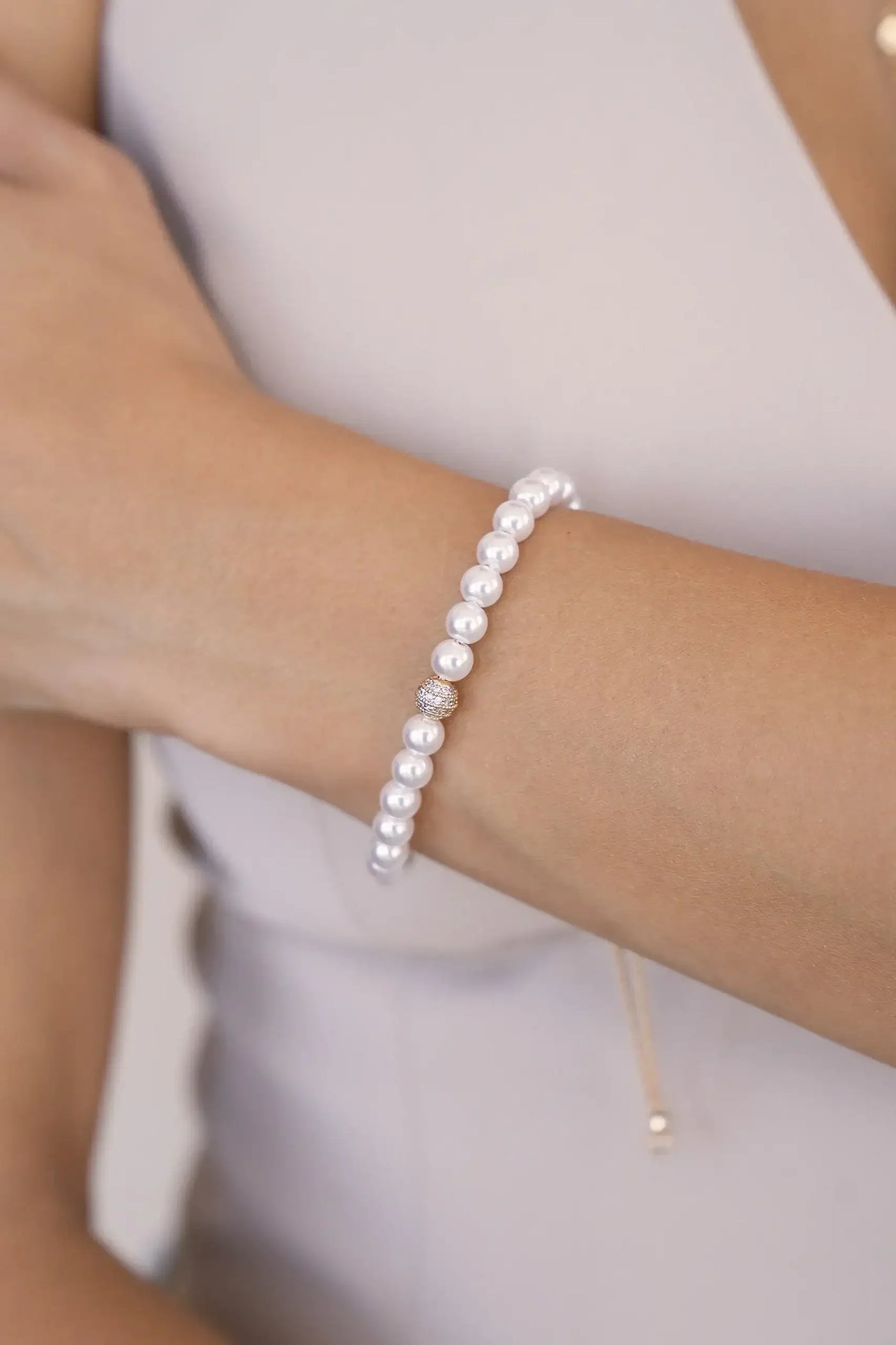 A pearl bracelet adorned with small circular pearls and a shiny centerpiece is worn on a hand with a light skin tone, against a neutral background with a soft, elegant attire.