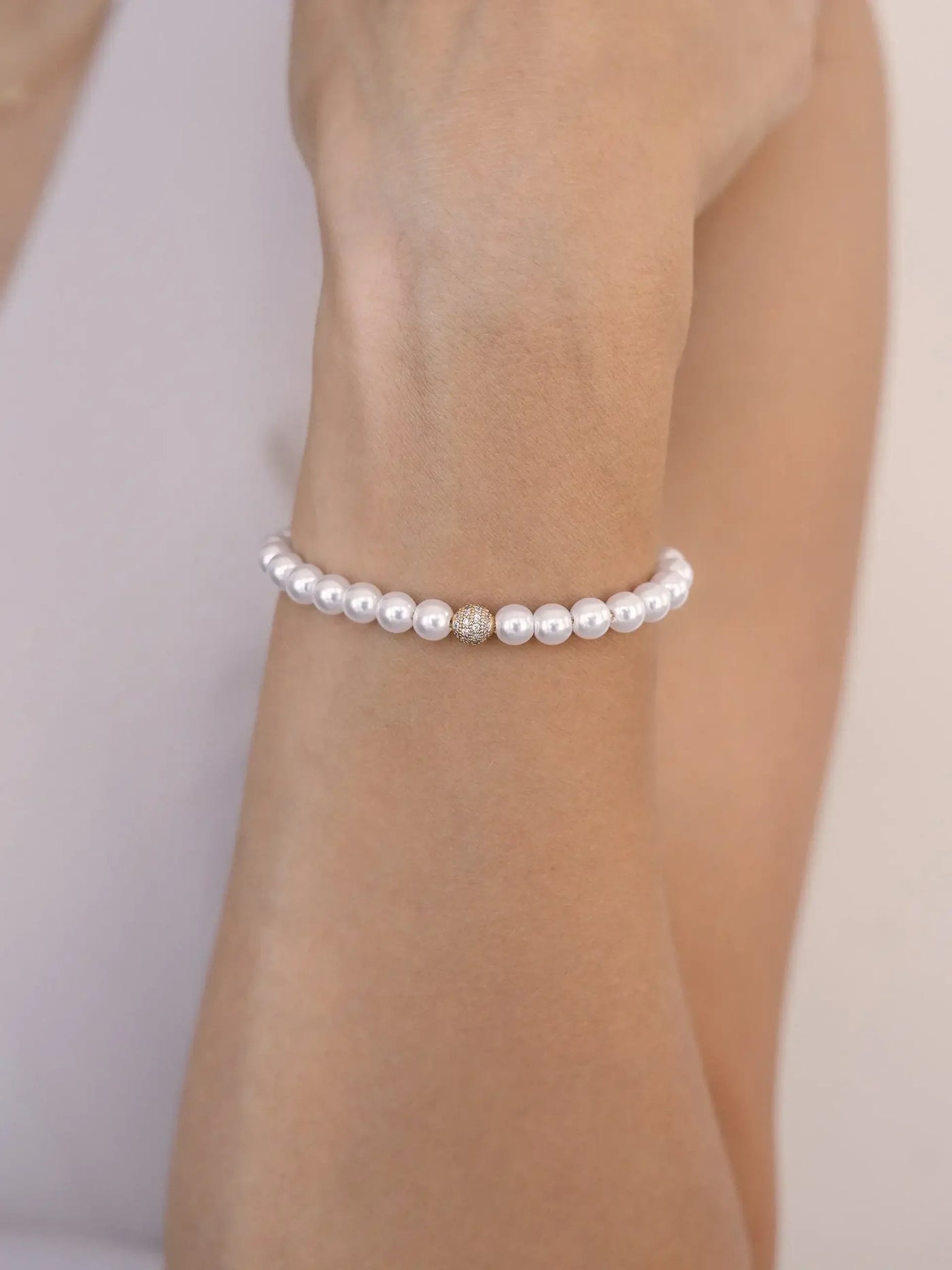 A delicate pearl bracelet adorns a wrist, featuring a central golden bead. The hand is positioned elegantly, with a neutral background enhancing the bracelet's simplicity and elegance.