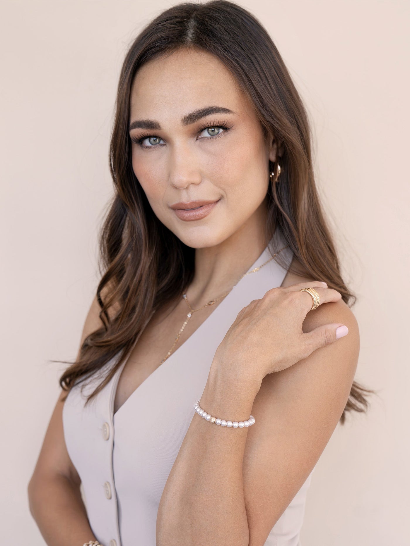 A woman with long, dark hair poses elegantly, touching her shoulder. She wears a light-colored sleeveless top and delicate jewelry. The background is a soft, neutral tone, enhancing her features.