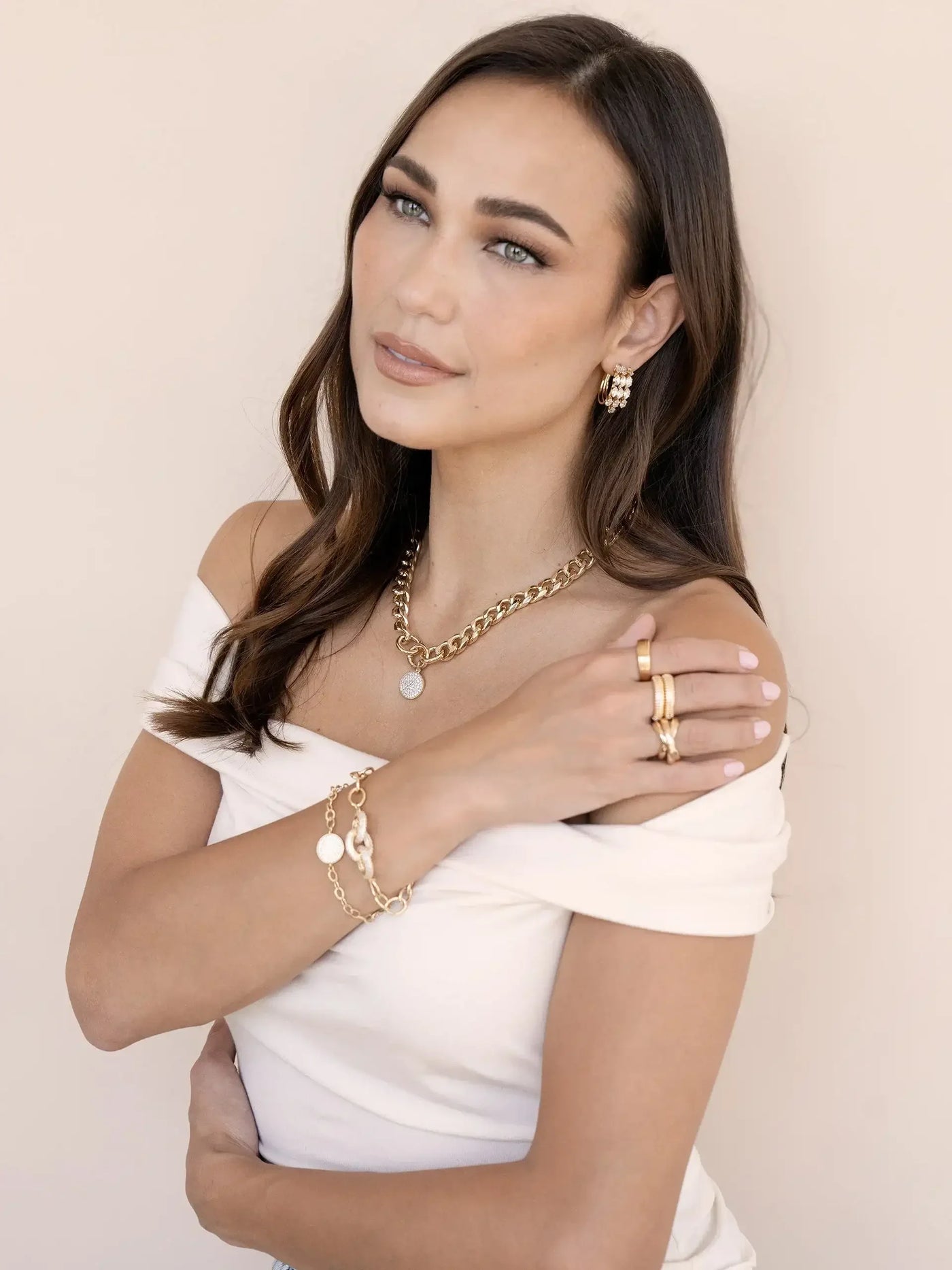 A woman with long, flowing hair poses gracefully, dressed in a white off-shoulder top adorned with various gold jewelry, set against a soft beige background.