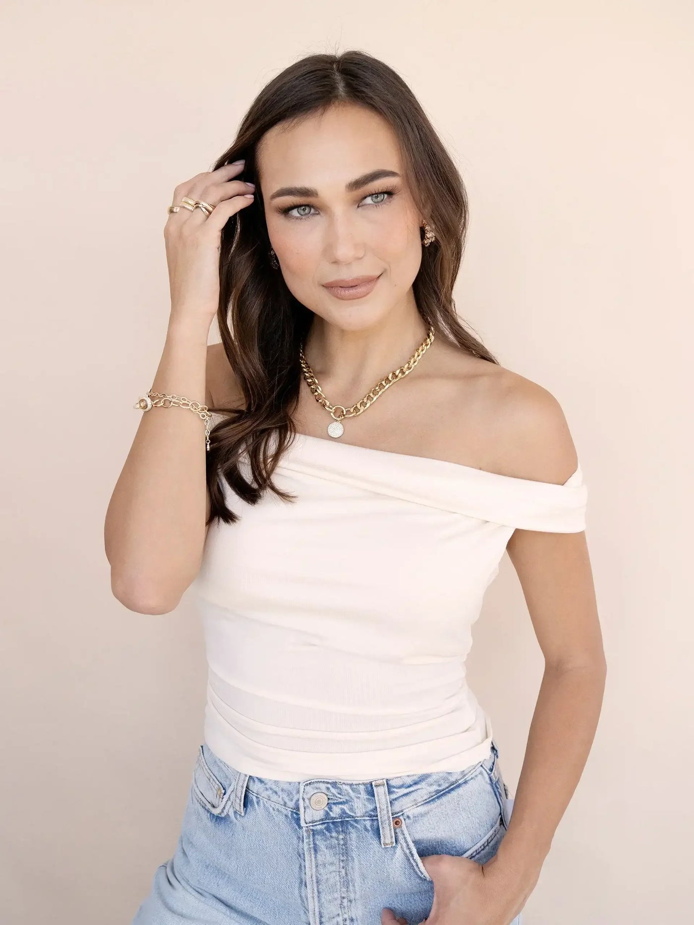 A woman with long brown hair wears an off-shoulder top and high-waisted denim shorts. She is touching her hair, smiling, against a soft, neutral background. Gold jewelry adorns her.
