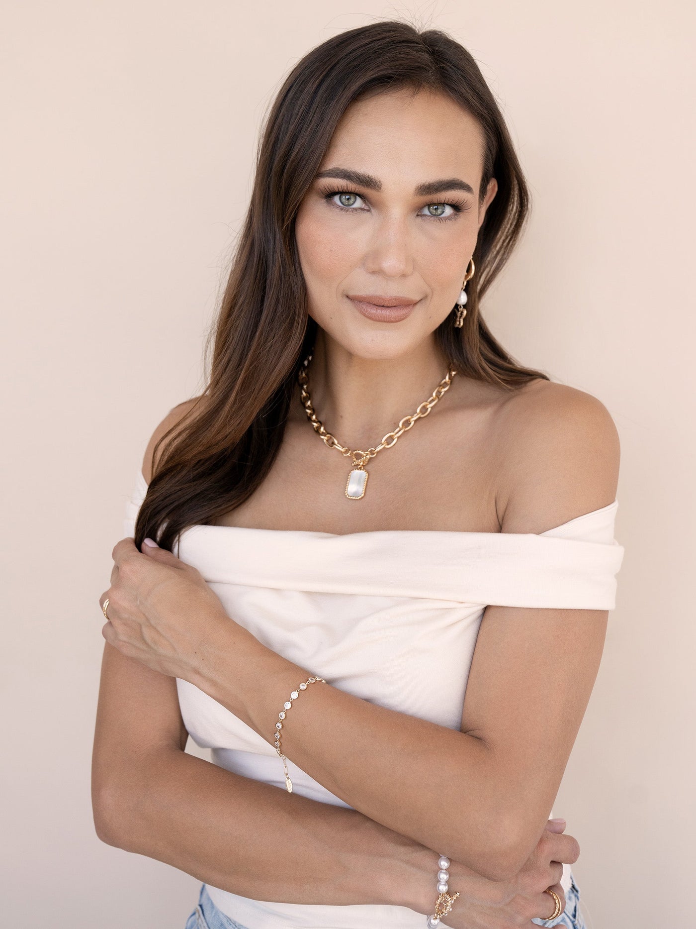 A woman with long brown hair poses gracefully, wearing an off-shoulder top and layered jewelry. She brings her arms around her torso, set against a soft beige background.