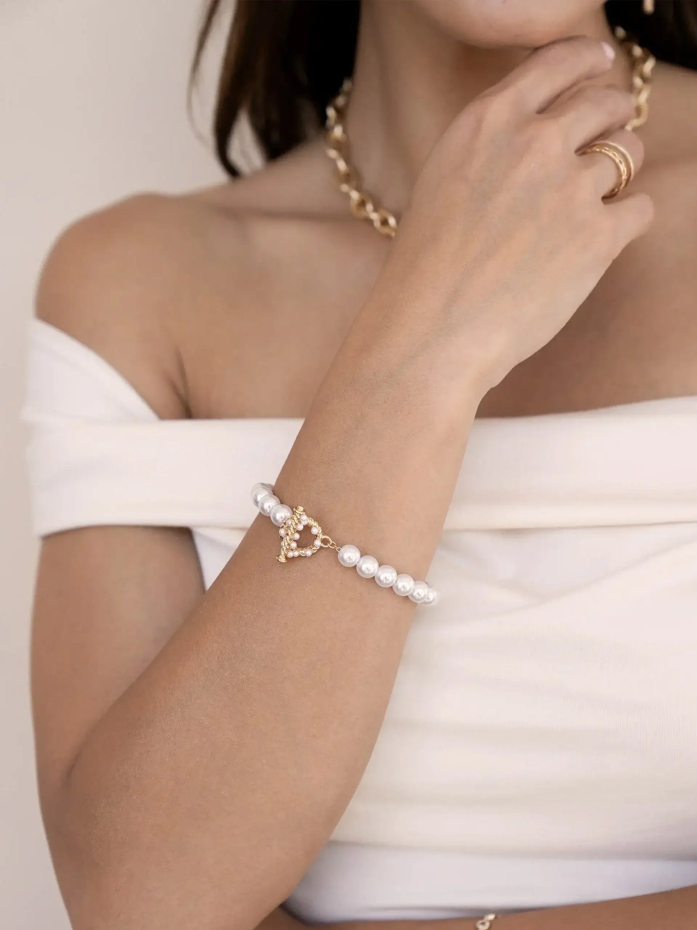 A hand adorned with a pearl bracelet showcases a heart-shaped charm, while the wearer lightly touches her neck. She is dressed in an off-the-shoulder white top, against a neutral backdrop.