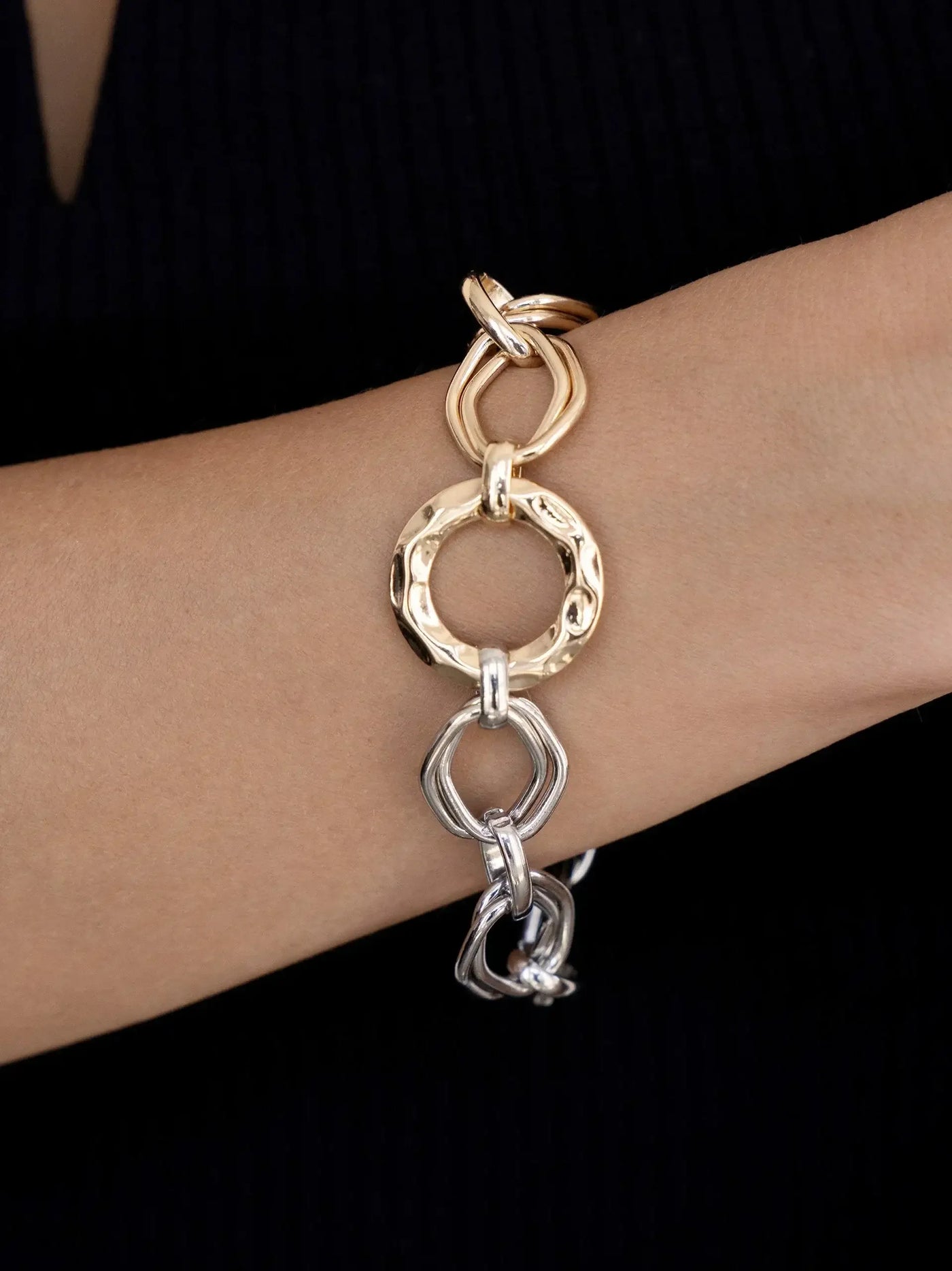 A metallic bracelet featuring interlinked gold and silver loops is worn on a wrist, showcasing a mix of textured circular and angular shapes against a dark, textured background.