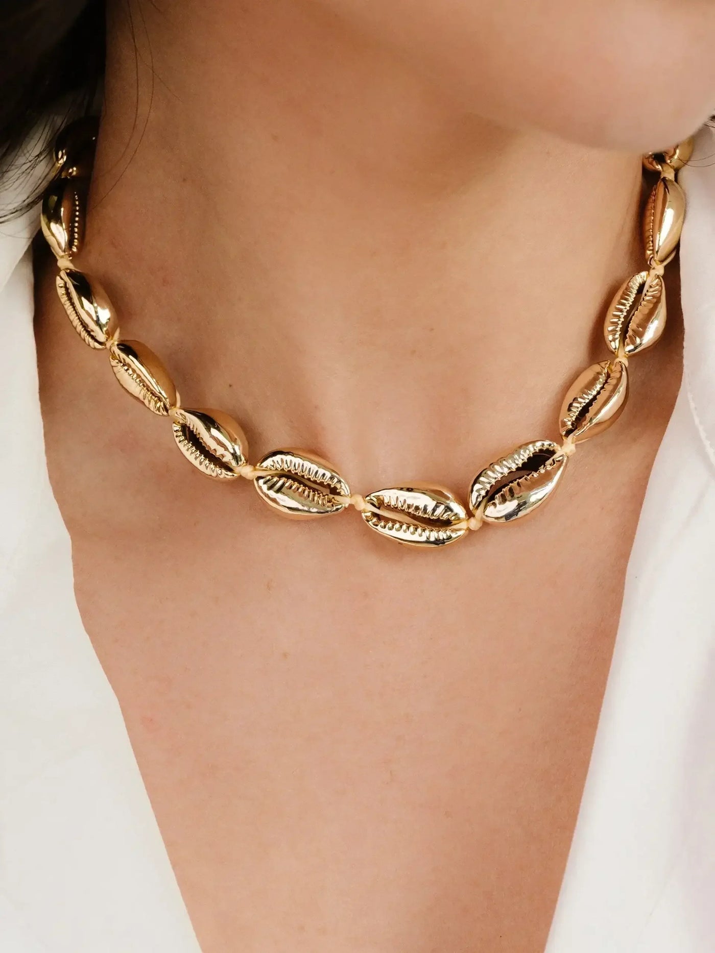 A gold-colored necklace with shell-like links adorns a person's neck. The wearer is dressed in a white top, against a softly lit, neutral background.