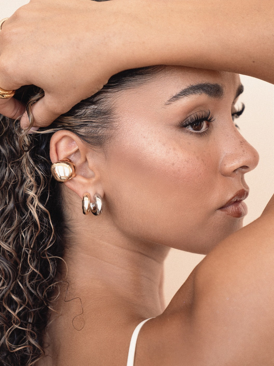 A woman with curly hair poses while touching her hair. She wears bold gold earrings and has a subtle, natural makeup look, set against a neutral background.