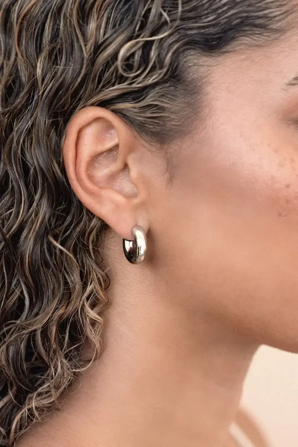 A close-up of a woman's ear adorned with a shiny silver hoop earring, with loose, wet, wavy hair cascading around her shoulder, emphasizing a natural and relaxed appearance.