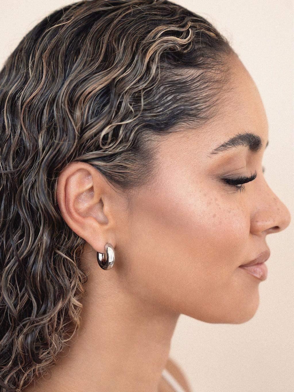A woman with curly hair is smiling softly, wearing a large silver hoop earring. The background features a smooth, light-colored surface, giving a warm and natural appearance.