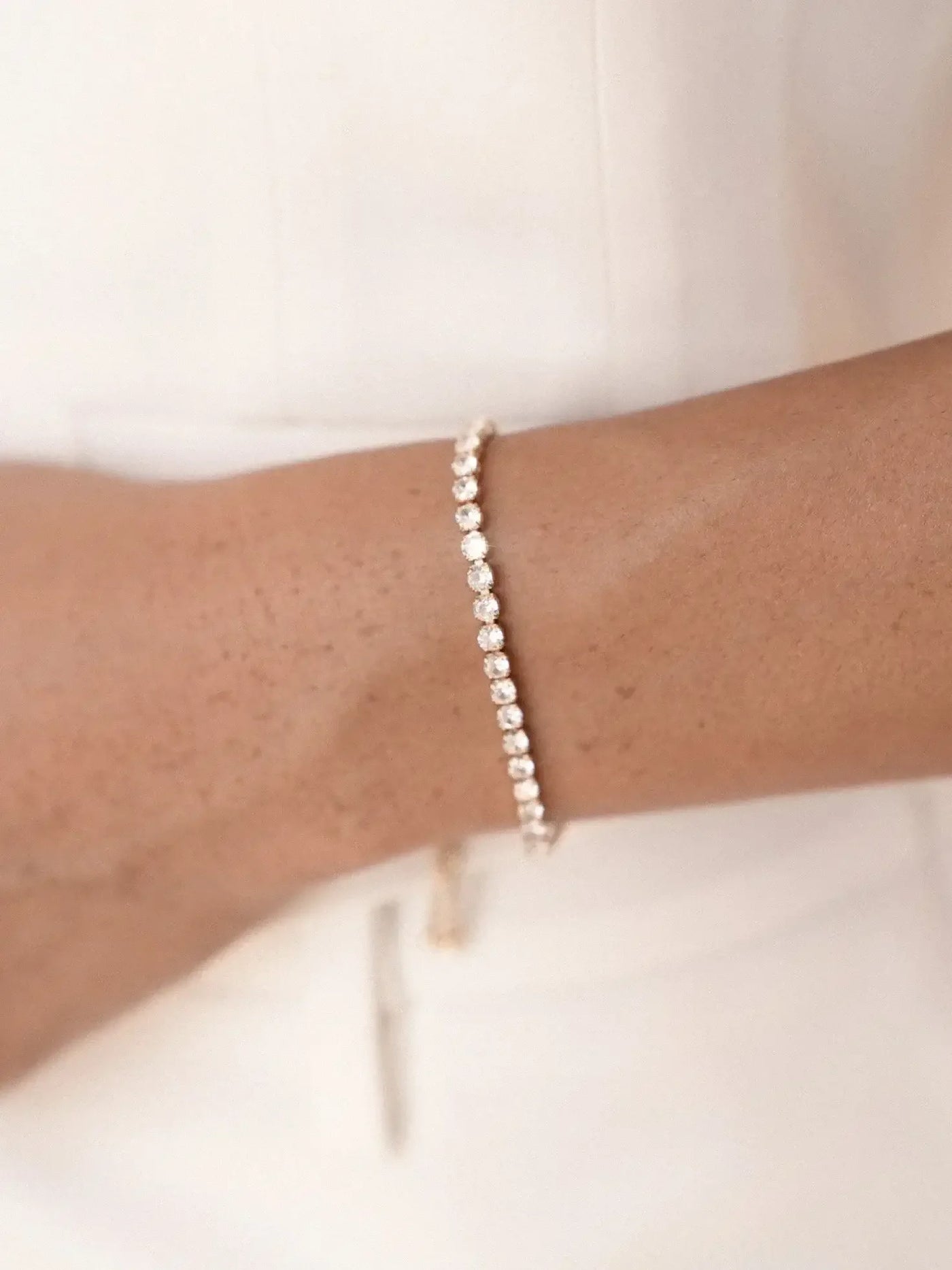A gold bracelet adorned with sparkling gemstones encircles a wrist. The skin tone is warm against the minimalist backdrop, emphasizing the elegance of the jewelry and the person’s attire.