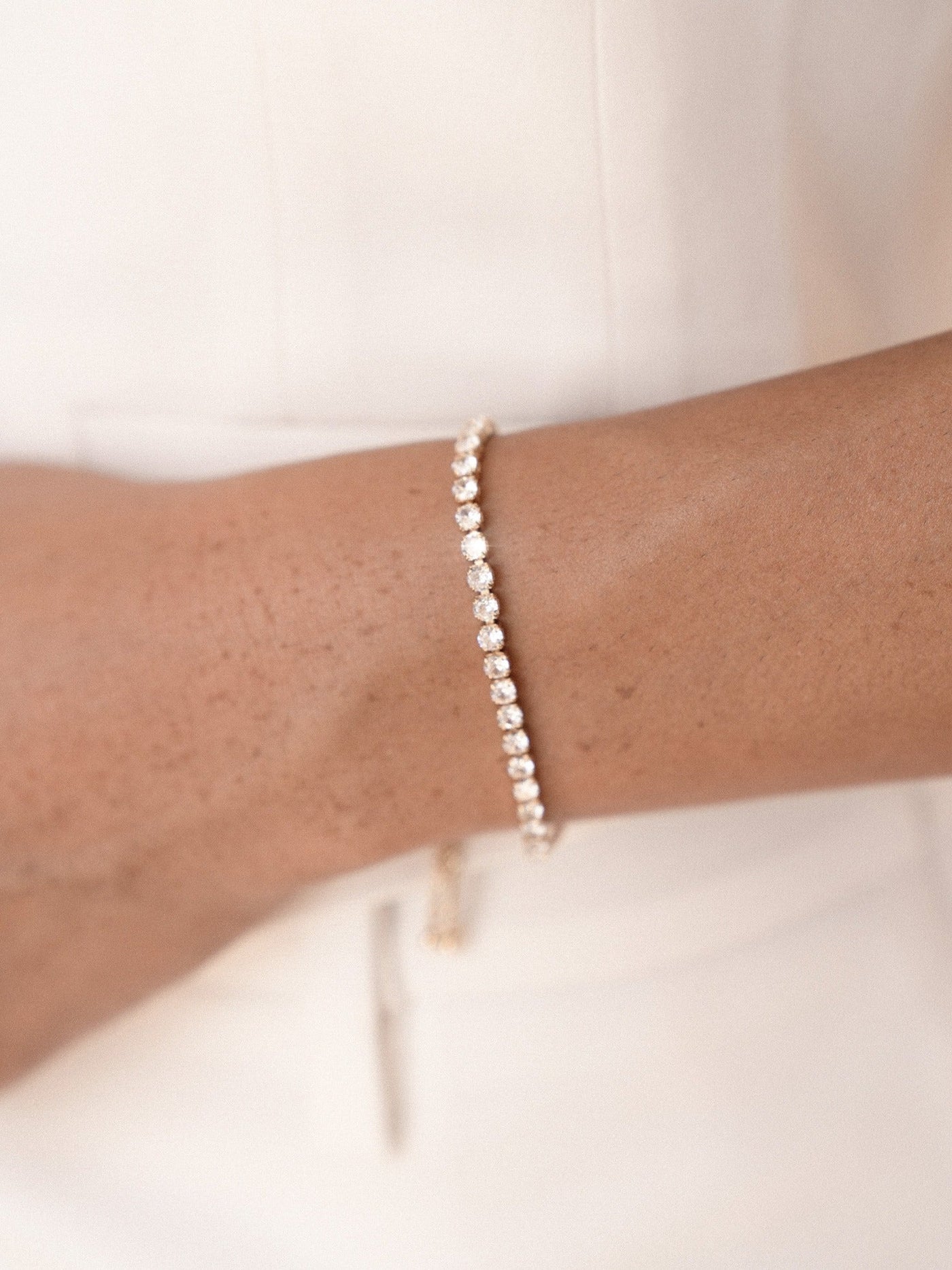 A gold bracelet adorned with sparkling gemstones encircles a wrist. The skin tone is warm against the minimalist backdrop, emphasizing the elegance of the jewelry and the person’s attire.