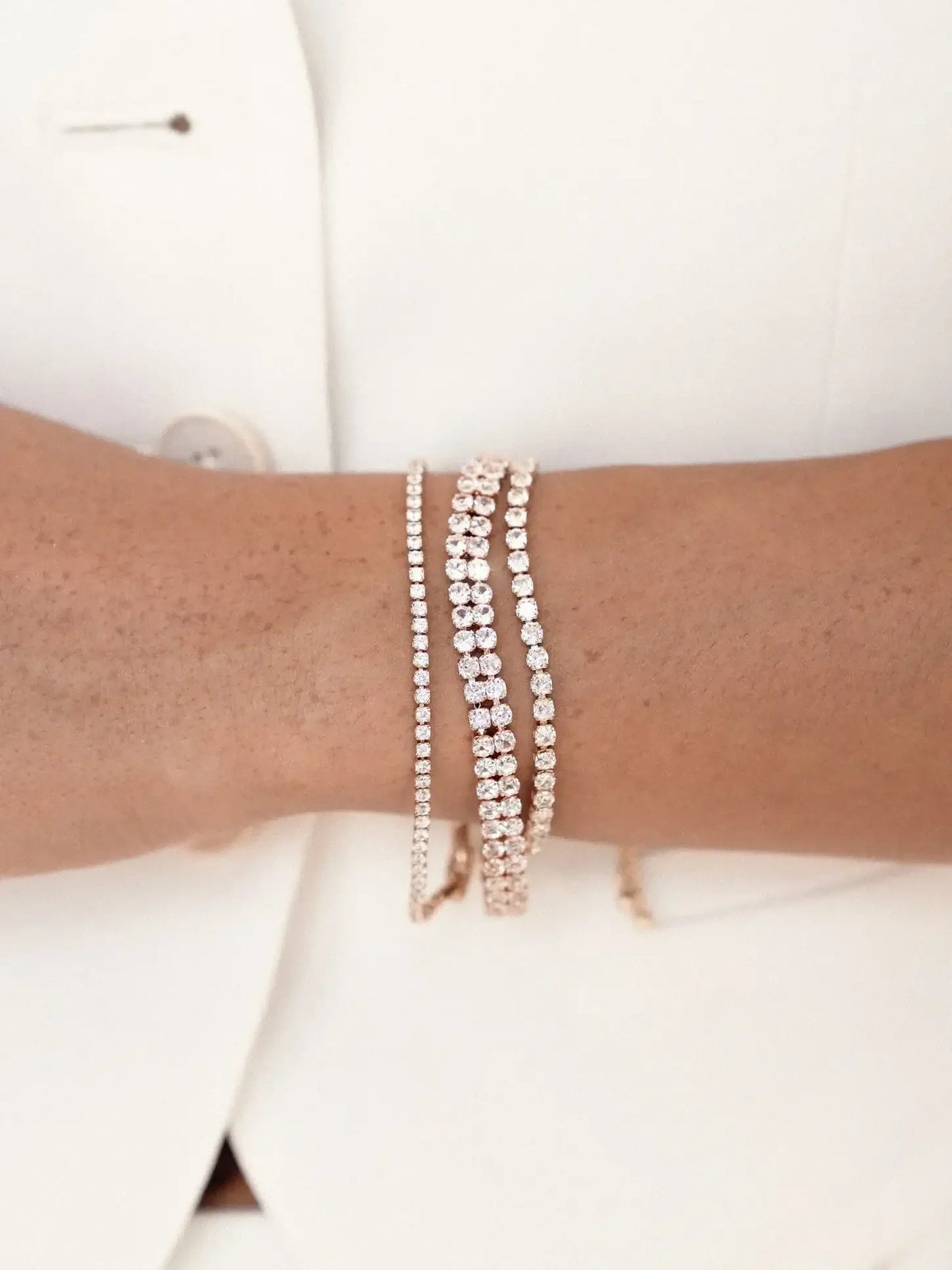 A wrist adorned with three shimmering gold bracelets, each featuring sparkling gemstones. The hand is resting against a neutral-toned fabric background, enhancing the jewelry's elegance.