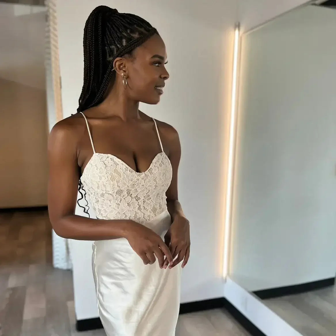 A woman stands in front of a mirror, wearing a stylish white dress with lace detailing. She smiles softly, posing confidently in a well-lit room with a neutral background.