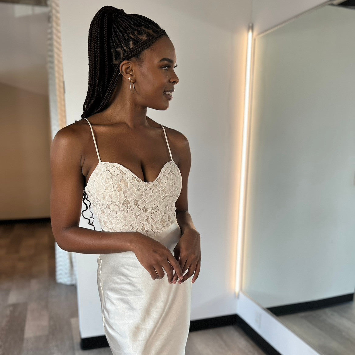 A woman stands in front of a mirror, wearing a stylish white dress with lace detailing. She smiles softly, posing confidently in a well-lit room with a neutral background.