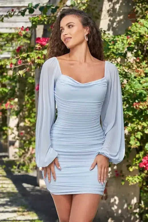 A woman stands confidently in a light blue dress with long, billowy sleeves, smiling softly. She is surrounded by lush greenery and vibrant pink flowers in a bright, sunny outdoor setting.