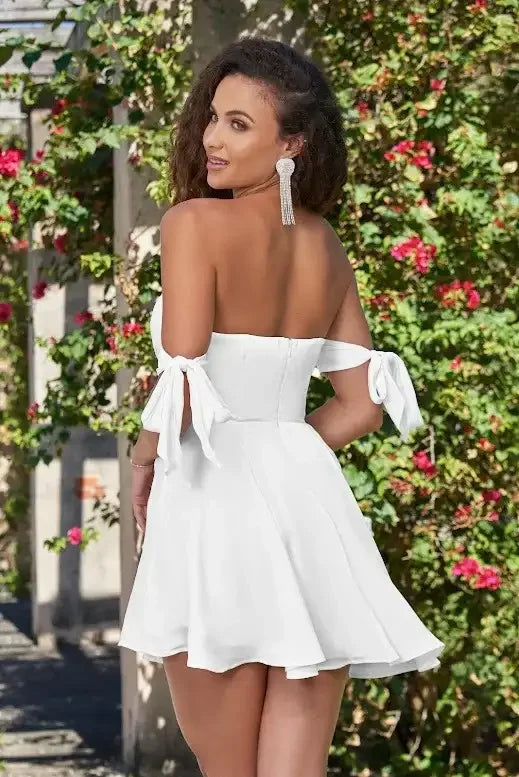 A woman wears a trendy white off-the-shoulder dress with bow ties on the sleeves, smiling over her shoulder amidst a backdrop of vibrant flowers and greenery.