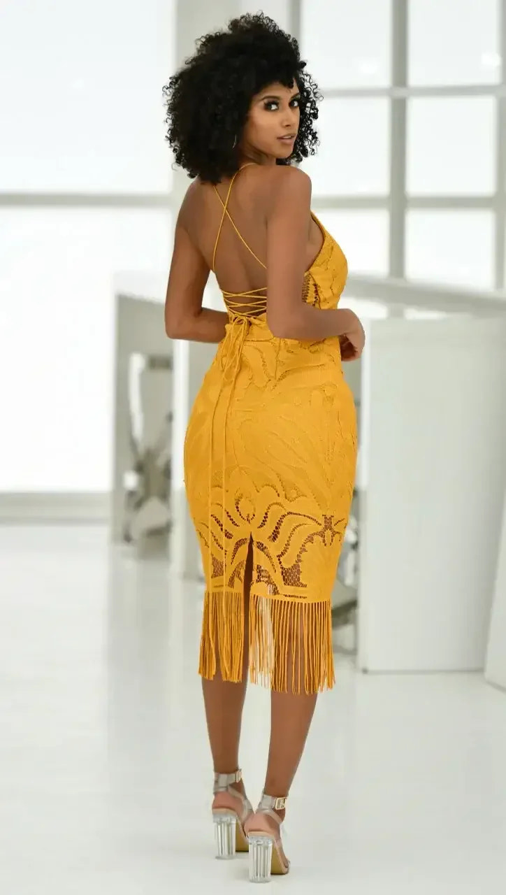 A woman with curly hair poses in a vibrant yellow, fringed dress that showcases intricate patterns. She stands in a bright, modern space with white walls and minimalistic decor.