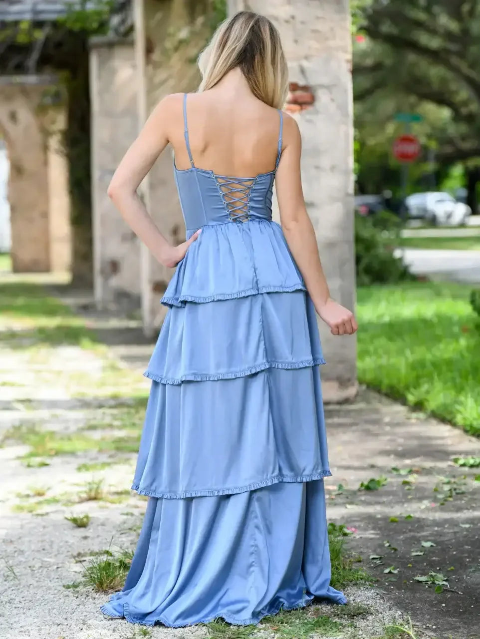 A light blue, tiered dress with spaghetti straps is worn by a woman, who stands with her back to the camera in a grassy area near a stone structure.