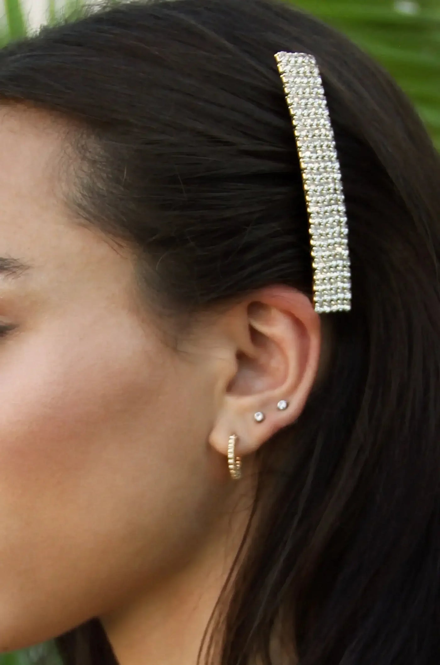A hair accessory adorned with rhinestones is secured in dark, wavy hair next to a woman’s ear, showcasing multiple earrings, while blurred greenery serves as the background.