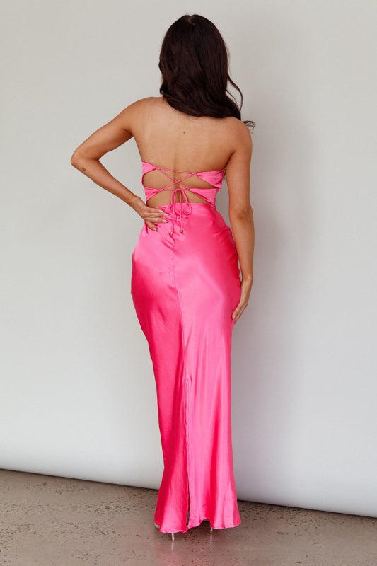A person wearing a long, pink satin dress stands with their back facing the viewer. The dress has a strapless design and ties intricately at the back, displaying a smooth, minimalist background.