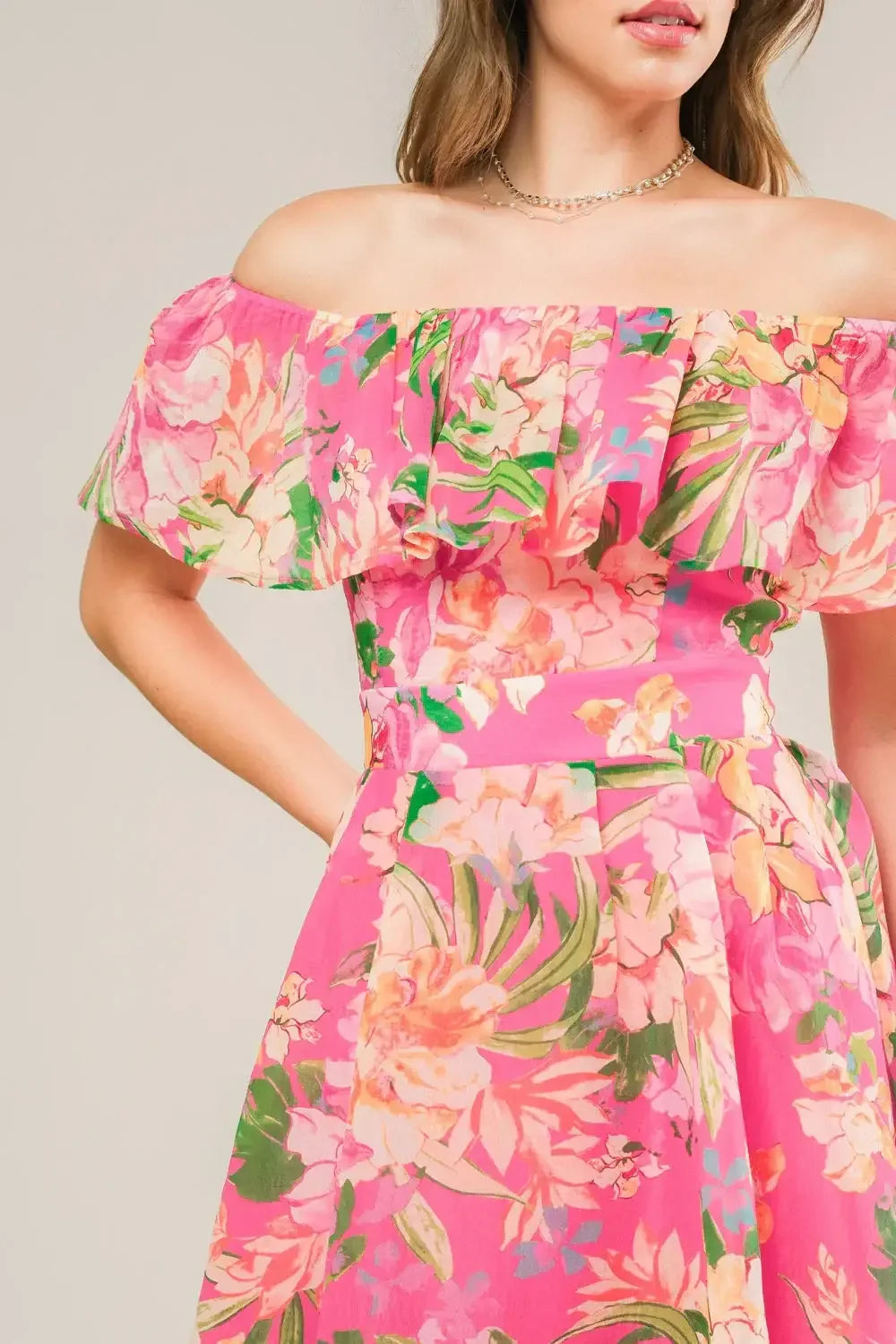 A floral dress in vibrant pink features an off-the-shoulder neckline and ruffled sleeves. The dress has a fitted waist and is set against a neutral background, highlighting its colorful design.