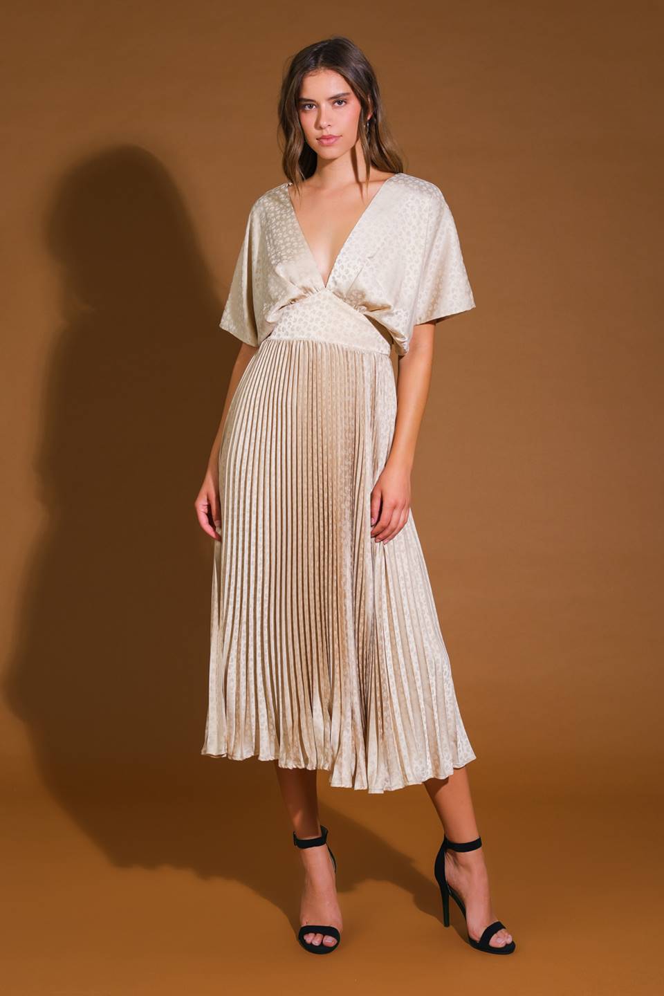 A woman stands confidently in a pleated, light-colored dress with a deep neckline and short sleeves, complemented by black high-heeled sandals. The backdrop is a warm, brown tone.