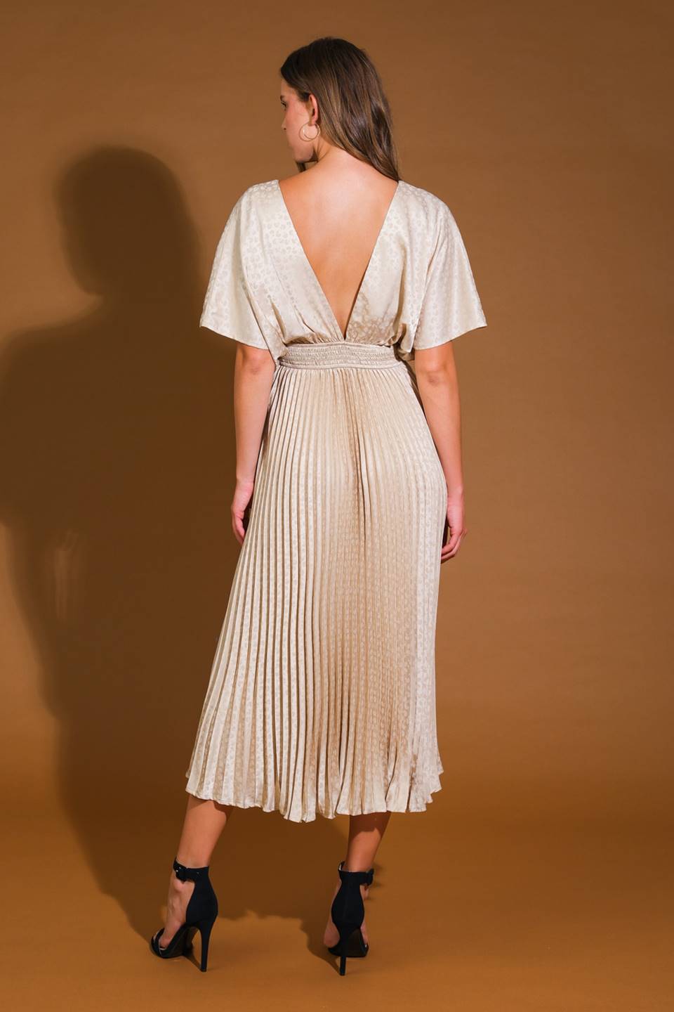 A woman stands with her back to the viewer, wearing a flowing, pleated, beige dress with a deep V-back. She is on a brown background, showcasing the dress's elegance.