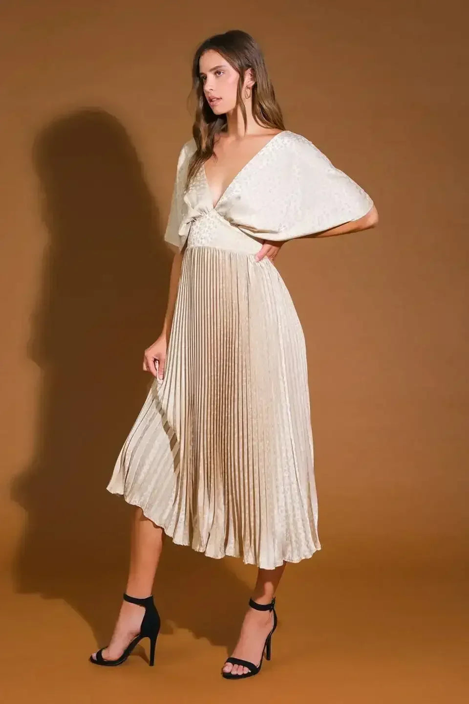 A woman wears a light, pleated dress with wide sleeves, striking a pose with one hand on her hip against a solid brown backdrop, complementing her look with black high heels.