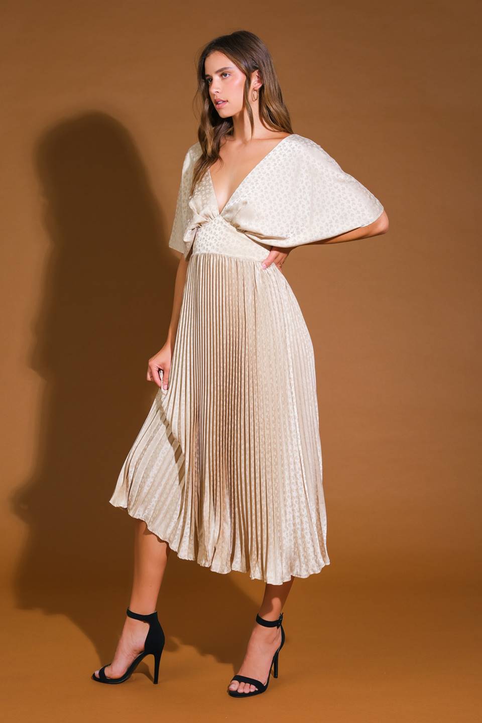 A woman wears a light, pleated dress with wide sleeves, striking a pose with one hand on her hip against a solid brown backdrop, complementing her look with black high heels.