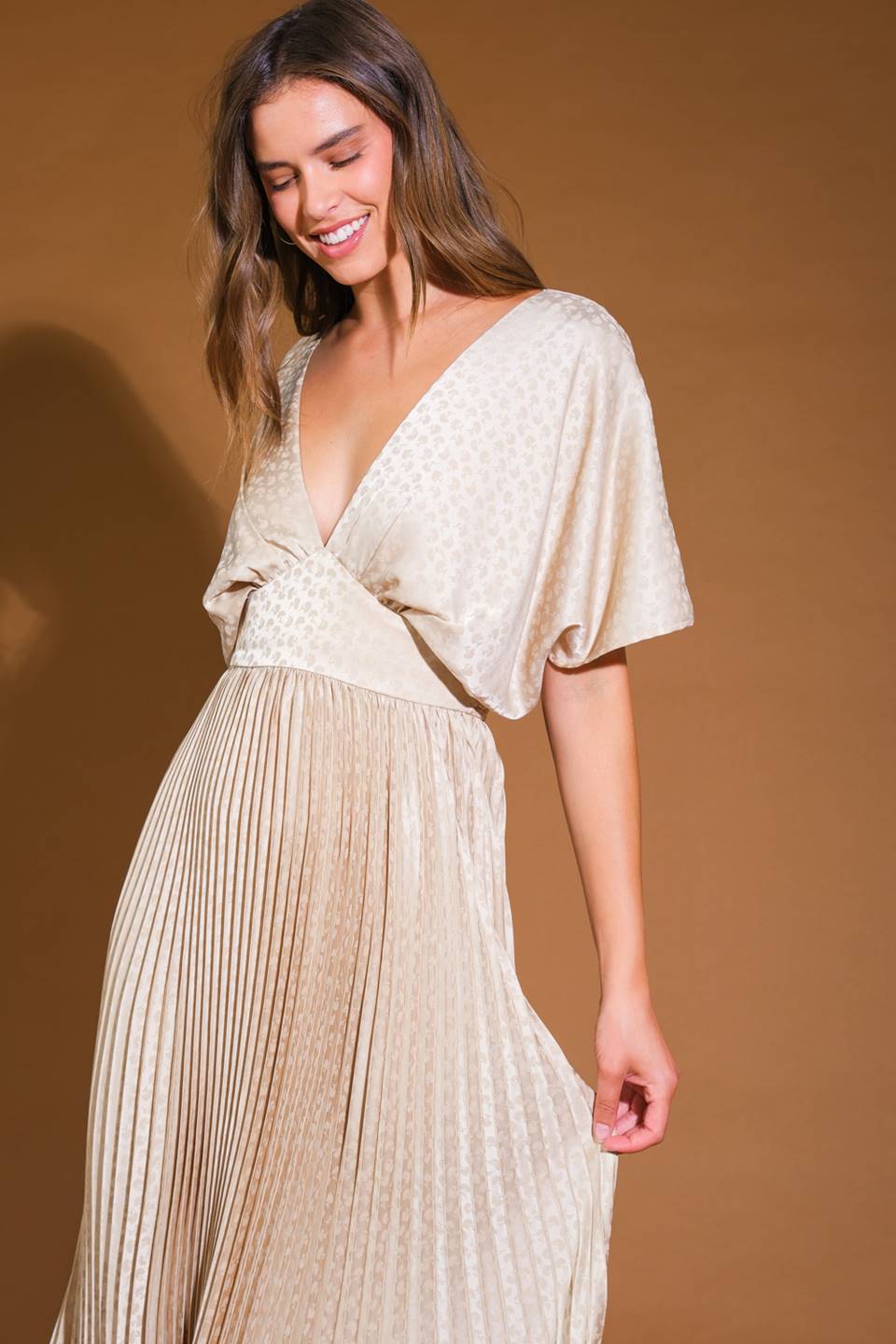 A woman wearing a flowing, light-colored dress with pleats smiles gently while tilting her head. The background is a soft, warm brown, enhancing the dress's delicate texture and design.