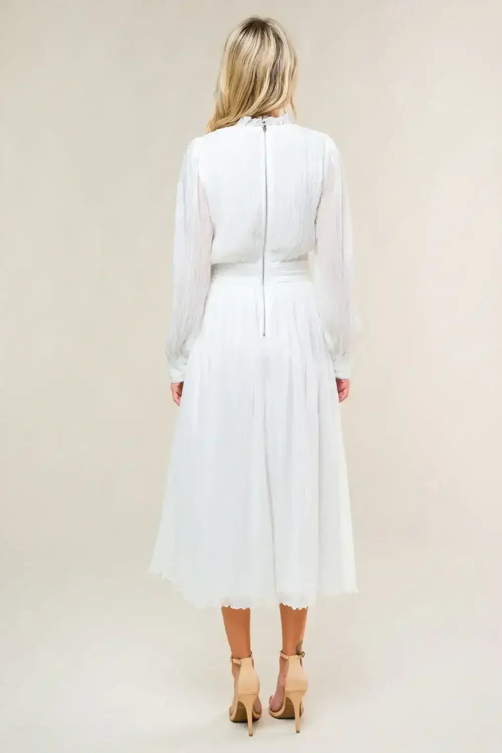A person in a white, flowy dress stands with their back visible, showcasing long sleeves and a high neckline, in a neutral-toned setting. The dress has a subtle texture.