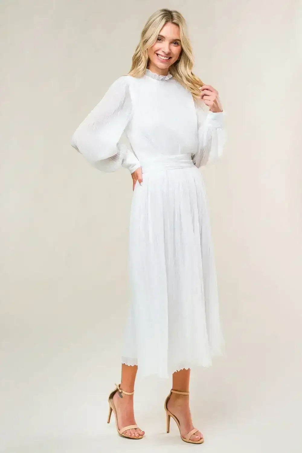 A woman in a flowing white dress with puffed sleeves smiles while standing on a light, neutral background. She wears strappy beige heels and is posed with one hand on her hip.