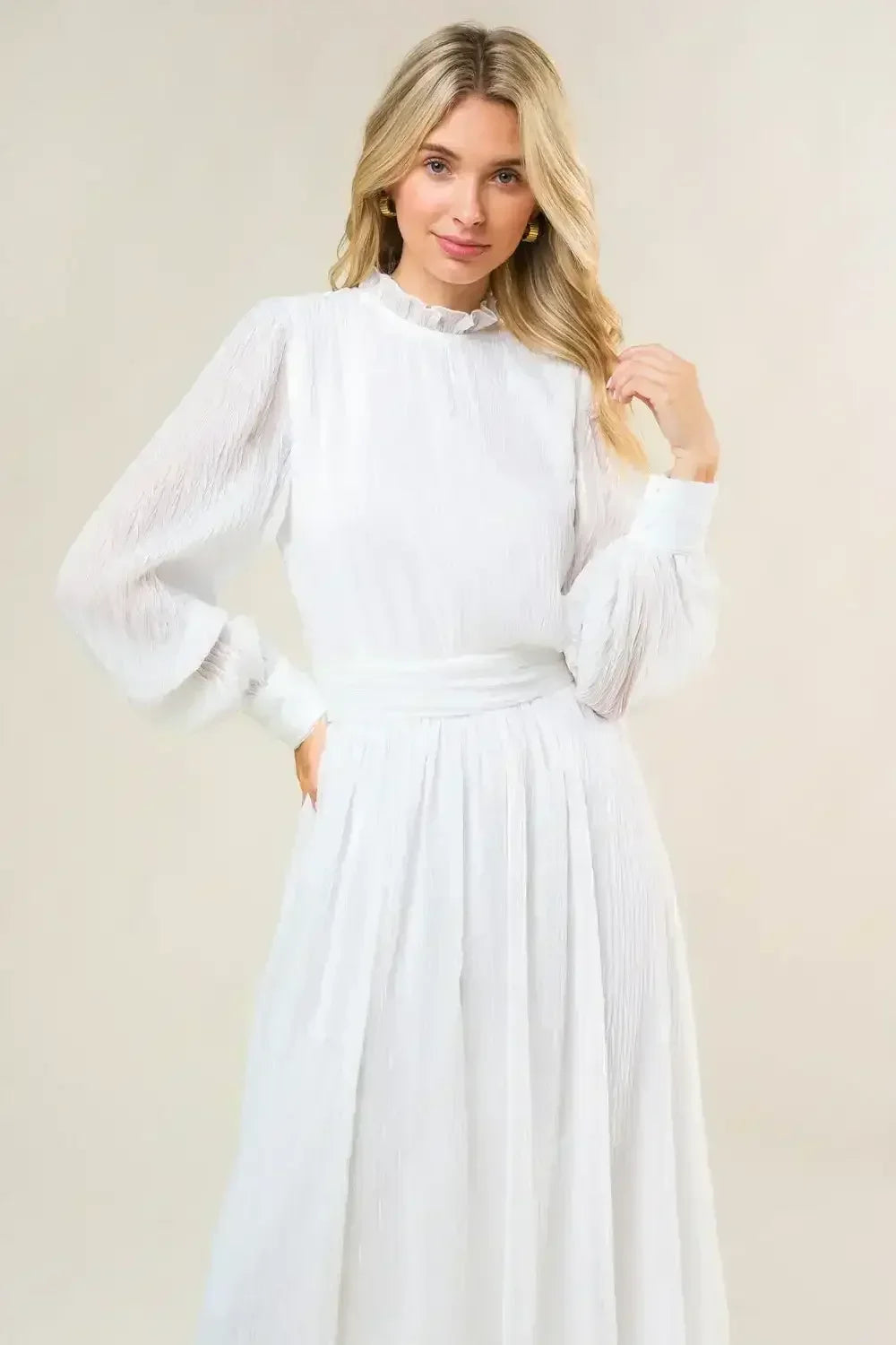 A woman wears a long, flowing white dress with a ruffled neckline and puffed sleeves, standing slightly angled and touching her hair, against a soft, neutral background.