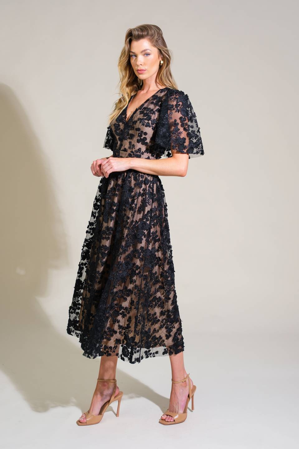 A woman stands elegantly in a sheer black floral dress, posing confidently. She wears nude high-heeled shoes, with a soft, neutral backdrop enhancing the outfit's delicate, intricate details.