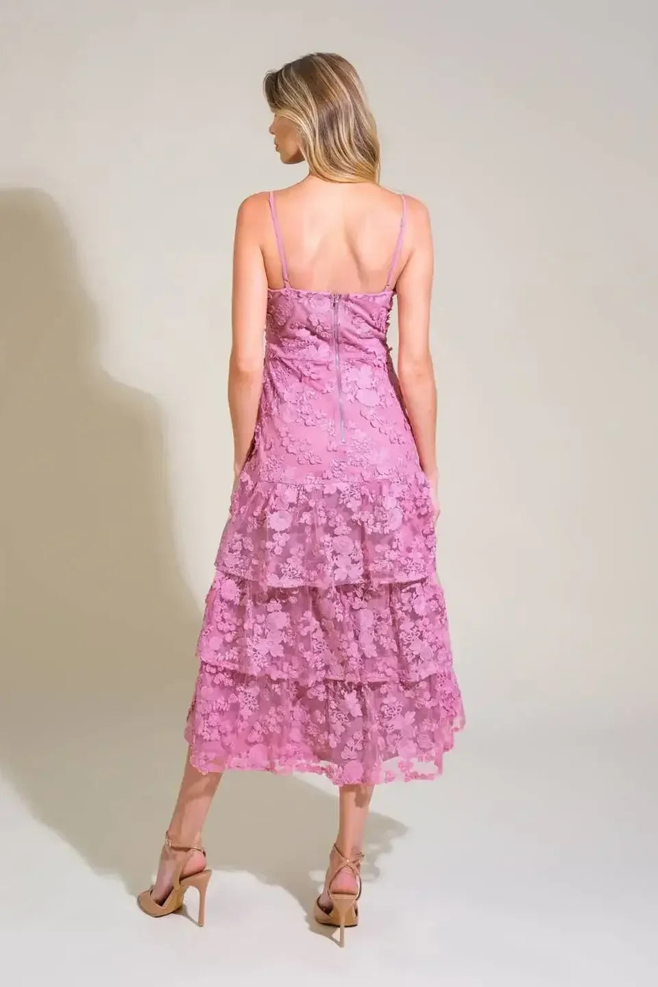 A woman stands with her back facing the viewer, wearing a tiered, floral lace pink dress and beige high-heeled sandals, against a neutral, softly lit background.