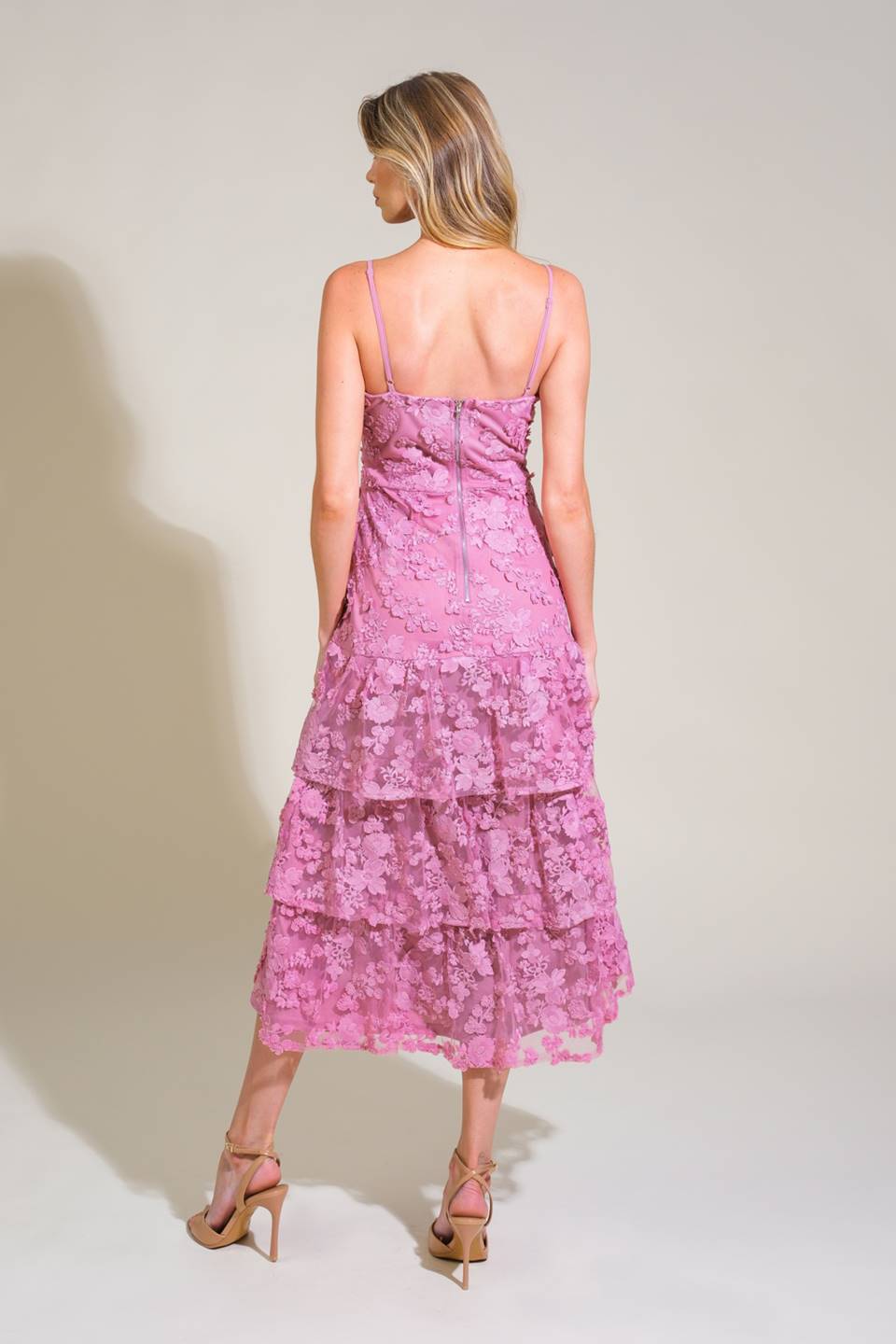 A woman stands with her back facing the viewer, wearing a tiered, floral lace pink dress and beige high-heeled sandals, against a neutral, softly lit background.