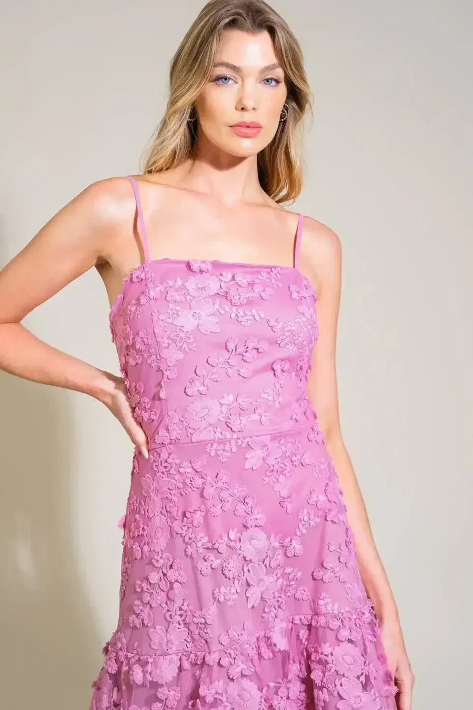 A model poses confidently in a pink, floral-embroidered dress with spaghetti straps, standing against a light beige background. She places one hand on her hip, showcasing the dress's delicate features.