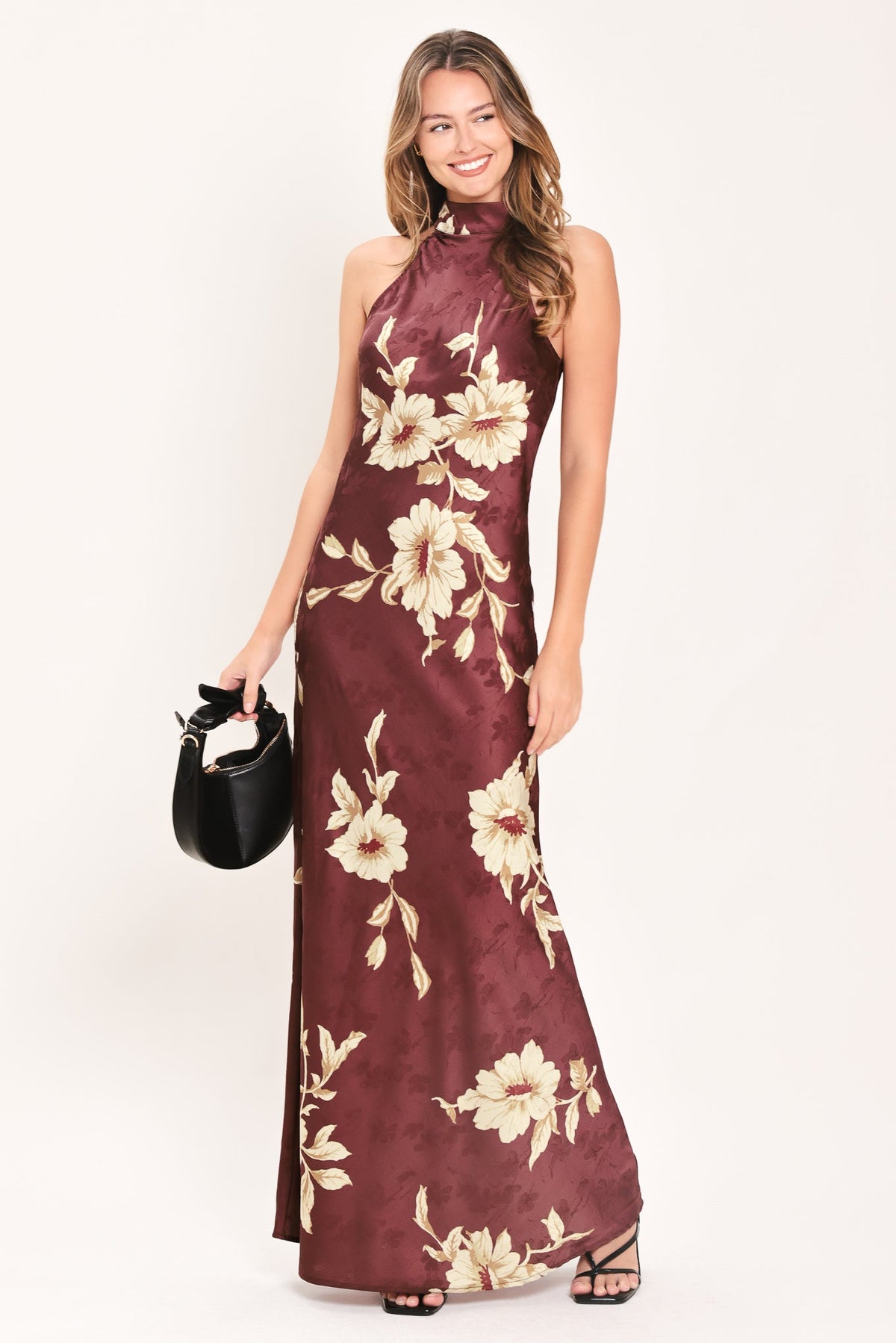 BORDEAUX IVORY BLOOM BURGUNDY MAXI DRESS | Lady Black Tie | A woman modeling a sleeveless, high-neck floor-length maxi dress in deep burgundy with a cream floral print, holding a small black handbag against a plain white studio background.