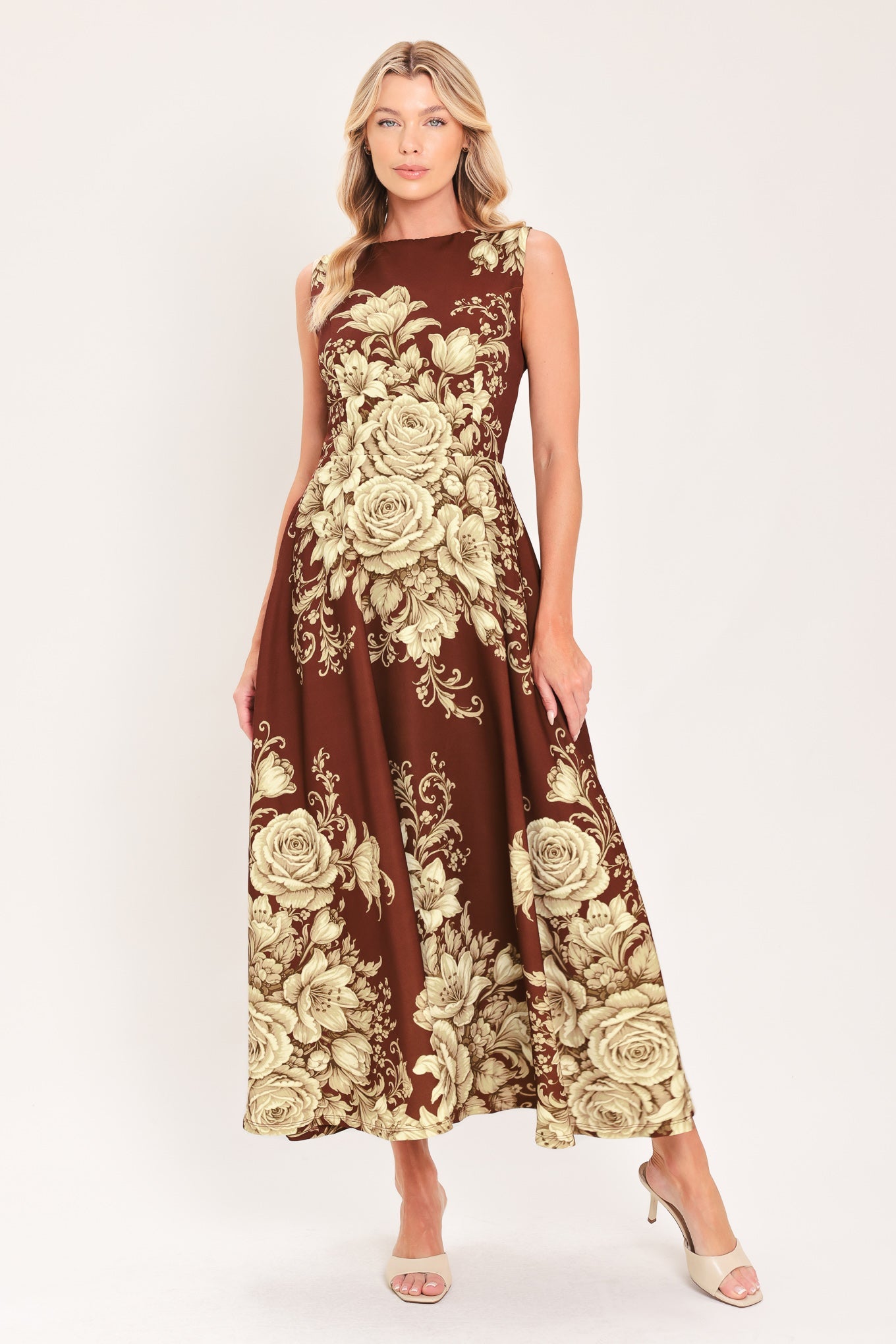 A woman modeling a sleeveless midi dress with a brown base and a large beige floral rose pattern, paired with beige high-heeled sandals, against a plain light background.