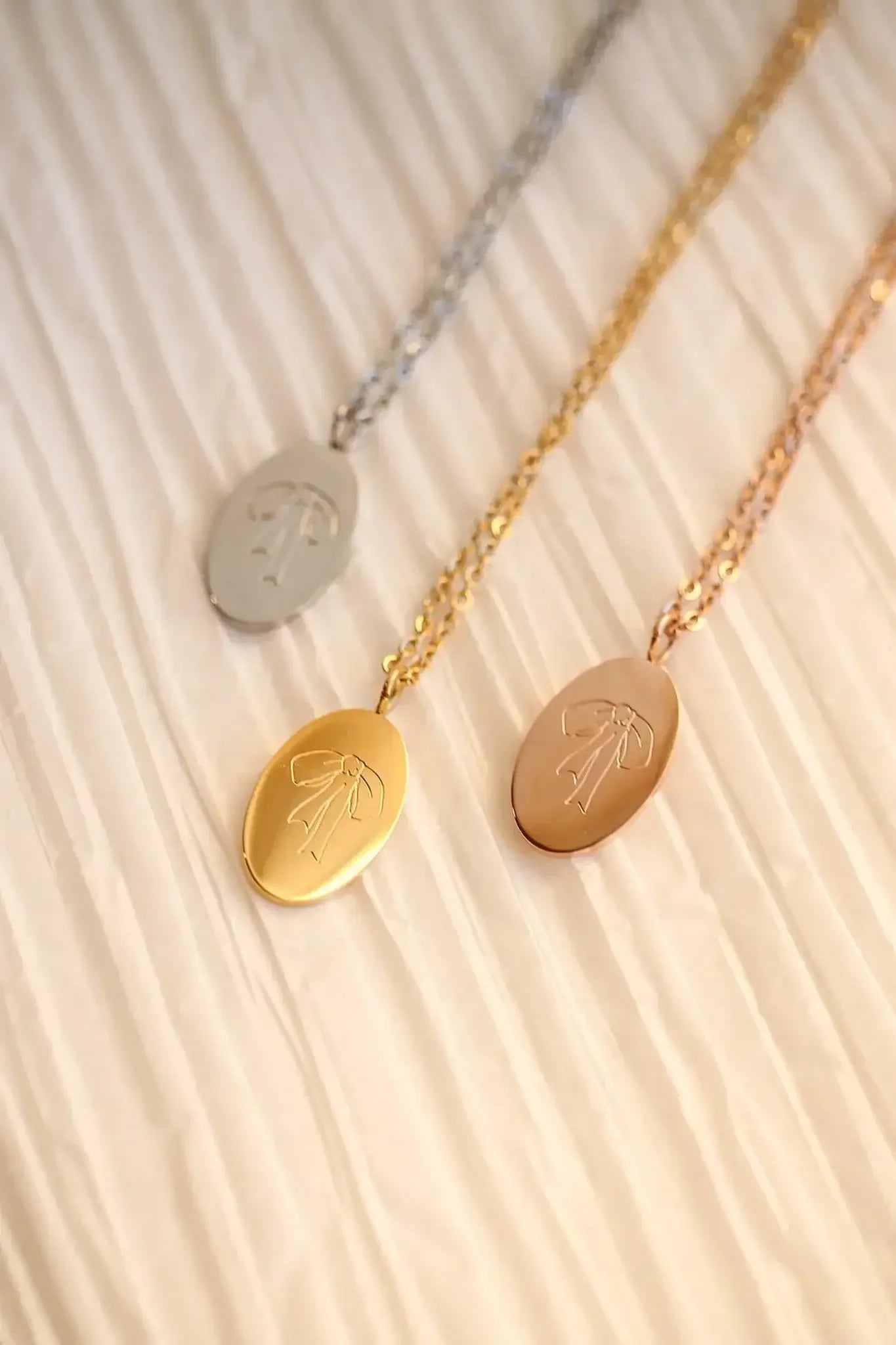 Three necklaces with oval pendants hang against a textured, light-colored background. Each pendant is a different metal: silver, gold, and rose gold, featuring an engraved bow design.