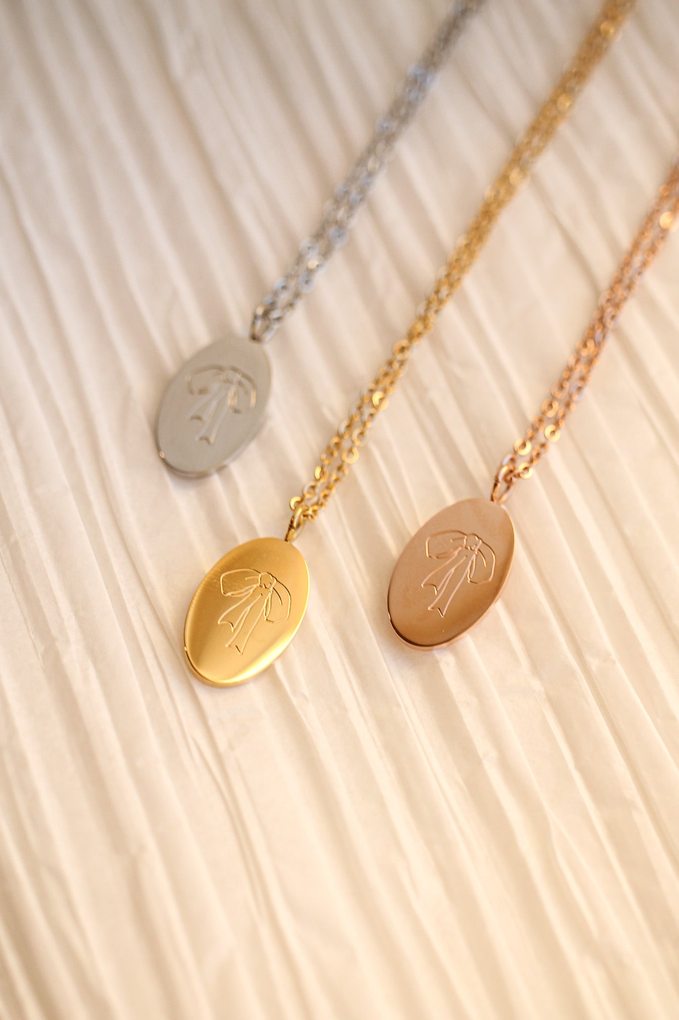 Three necklaces with oval pendants hang against a textured, light-colored background. Each pendant is a different metal: silver, gold, and rose gold, featuring an engraved bow design.