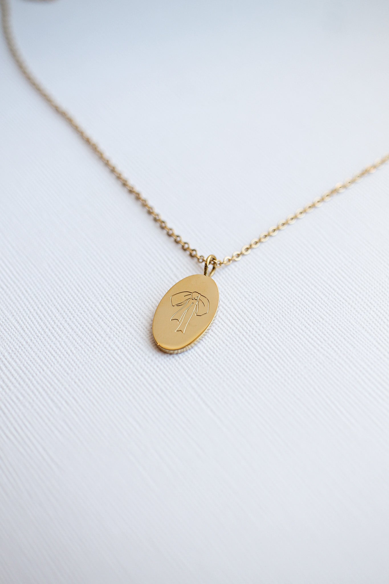 A gold necklace with an oval pendant lies on a textured white surface. The pendant features an etched design of a bow, reflecting a simple yet elegant style.