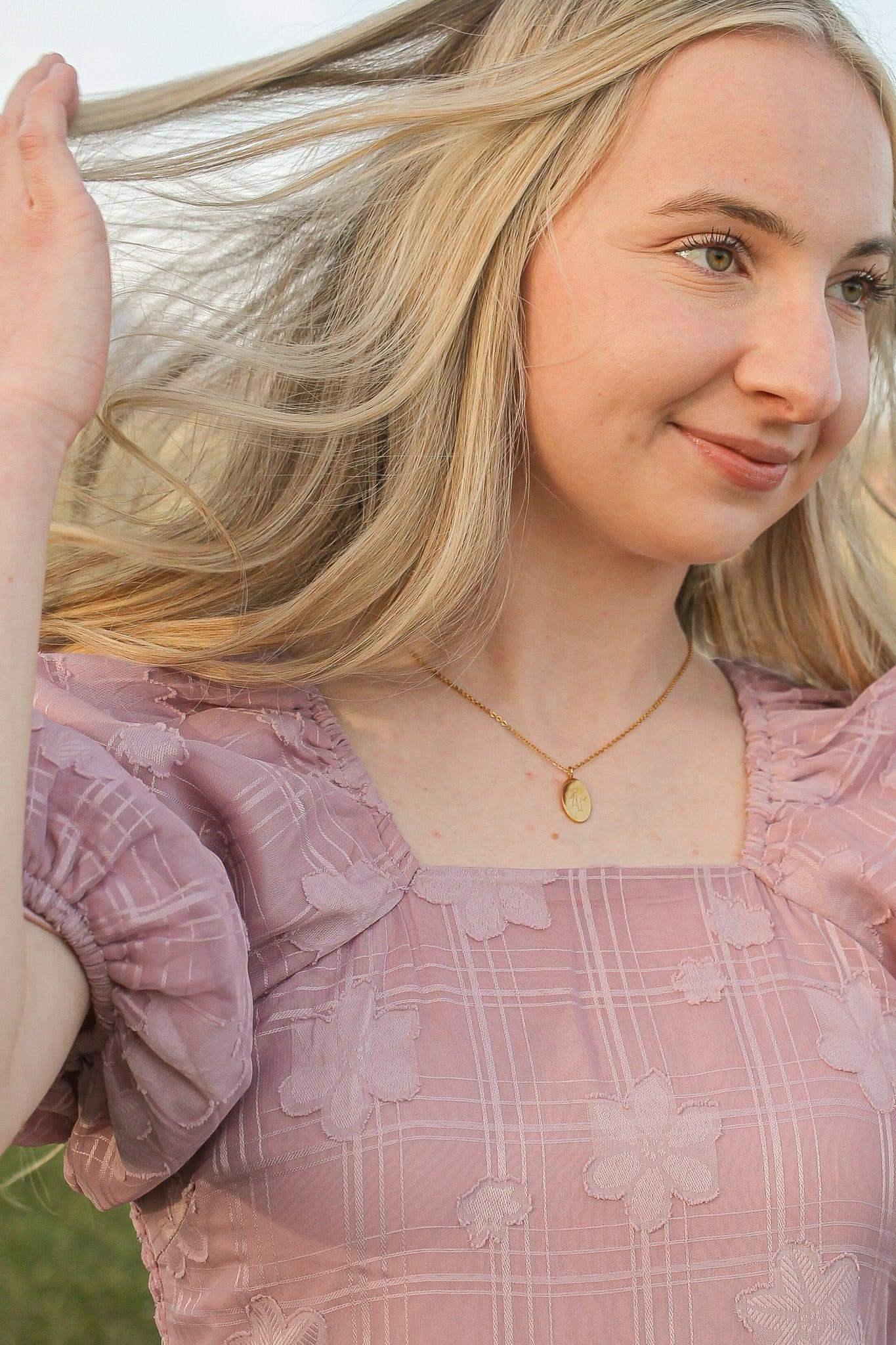 A young woman with long, flowing blonde hair gently tosses her hair back. She wears a light pink, floral-patterned dress and a gold necklace, set against a blurred, natural background.