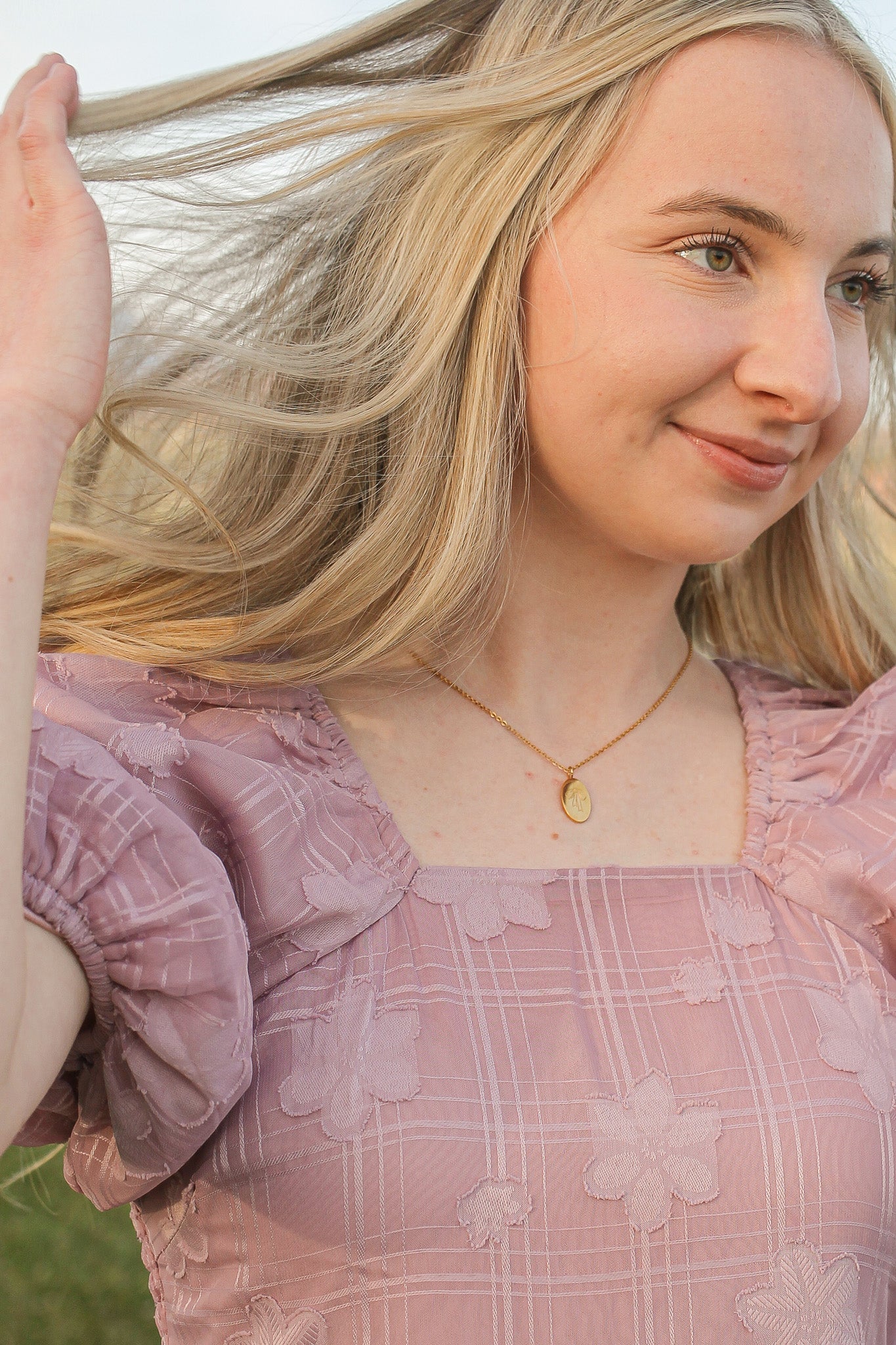 A young woman with long, flowing blonde hair gently tosses her hair back. She wears a light pink, floral-patterned dress and a gold necklace, set against a blurred, natural background.