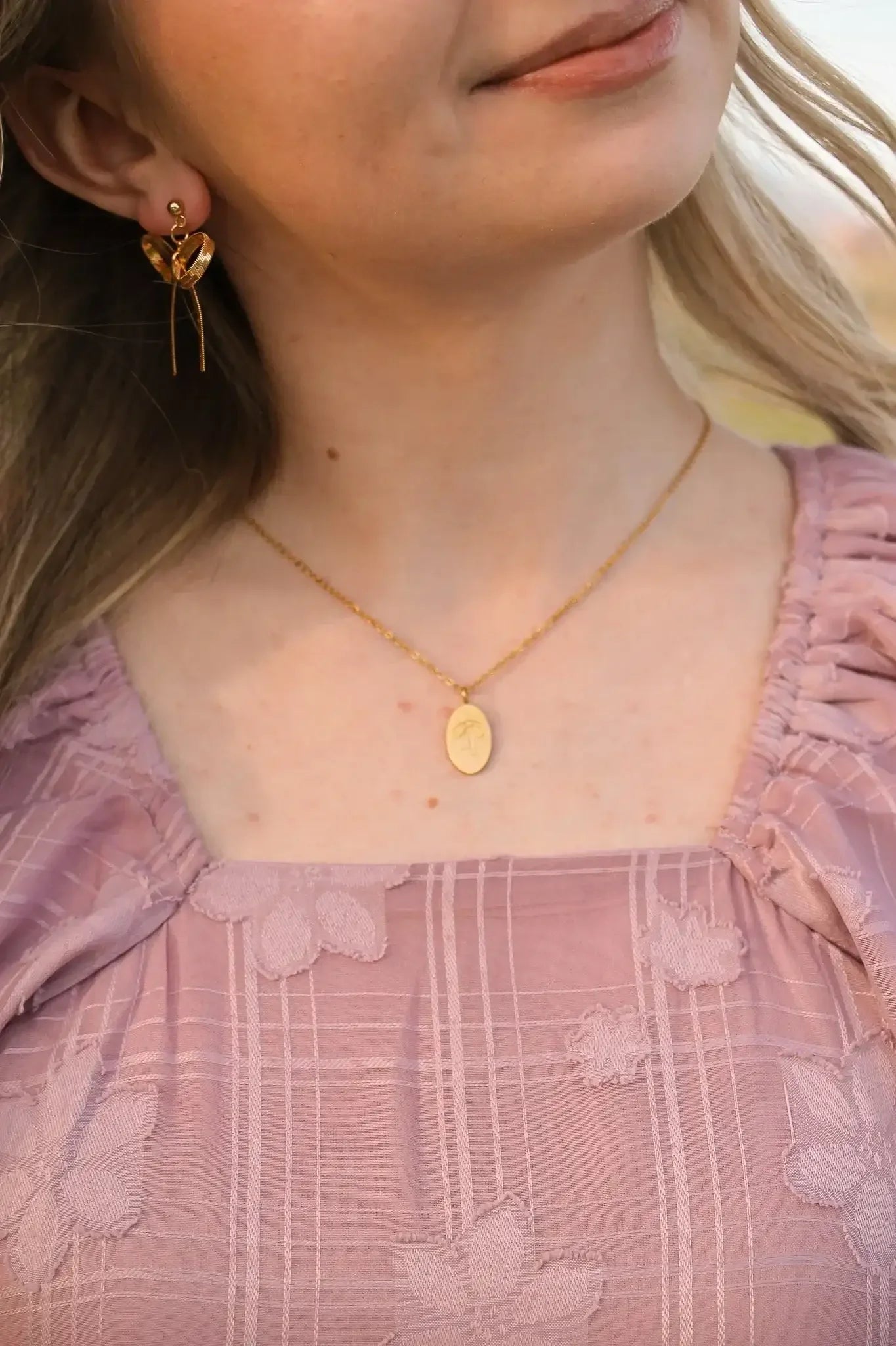 A young woman wears a floral-patterned lavender dress, adorned with gold earrings and a delicate necklace featuring a pendant, smiling softly in a natural outdoor setting.