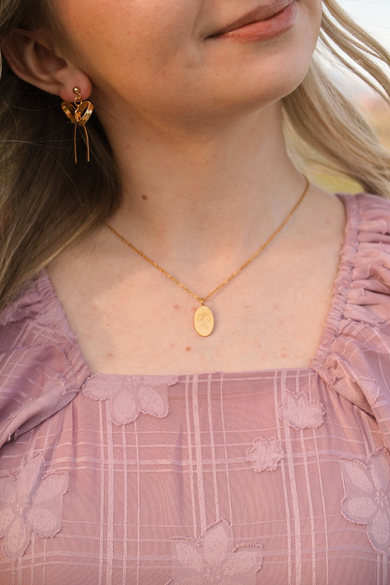 A young woman wears a floral-patterned lavender dress, adorned with gold earrings and a delicate necklace featuring a pendant, smiling softly in a natural outdoor setting.