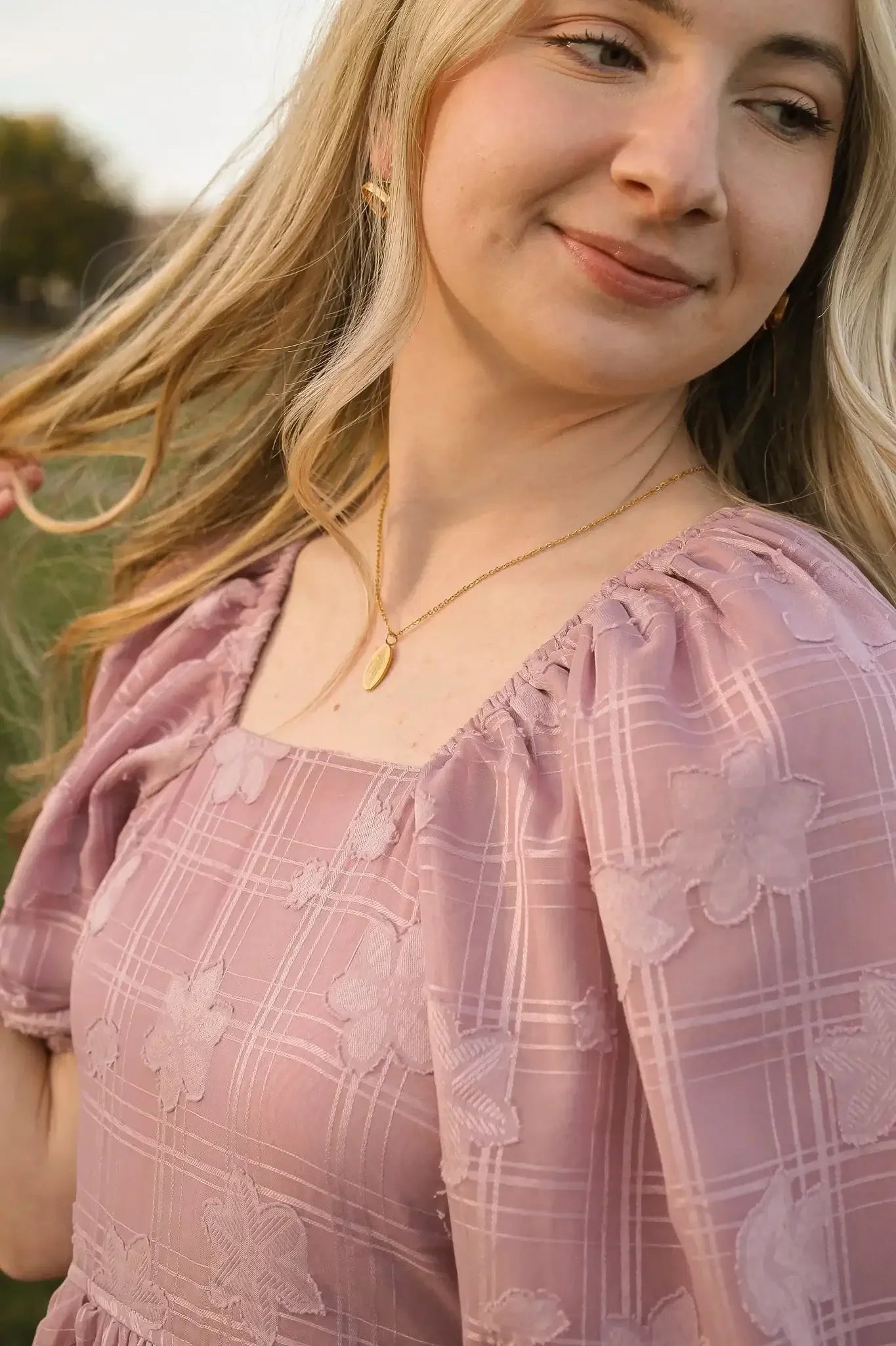 A young woman with long, wavy blonde hair is gently playing with her hair while wearing a floral-patterned, light purple top. The background hints at a natural, outdoor setting.