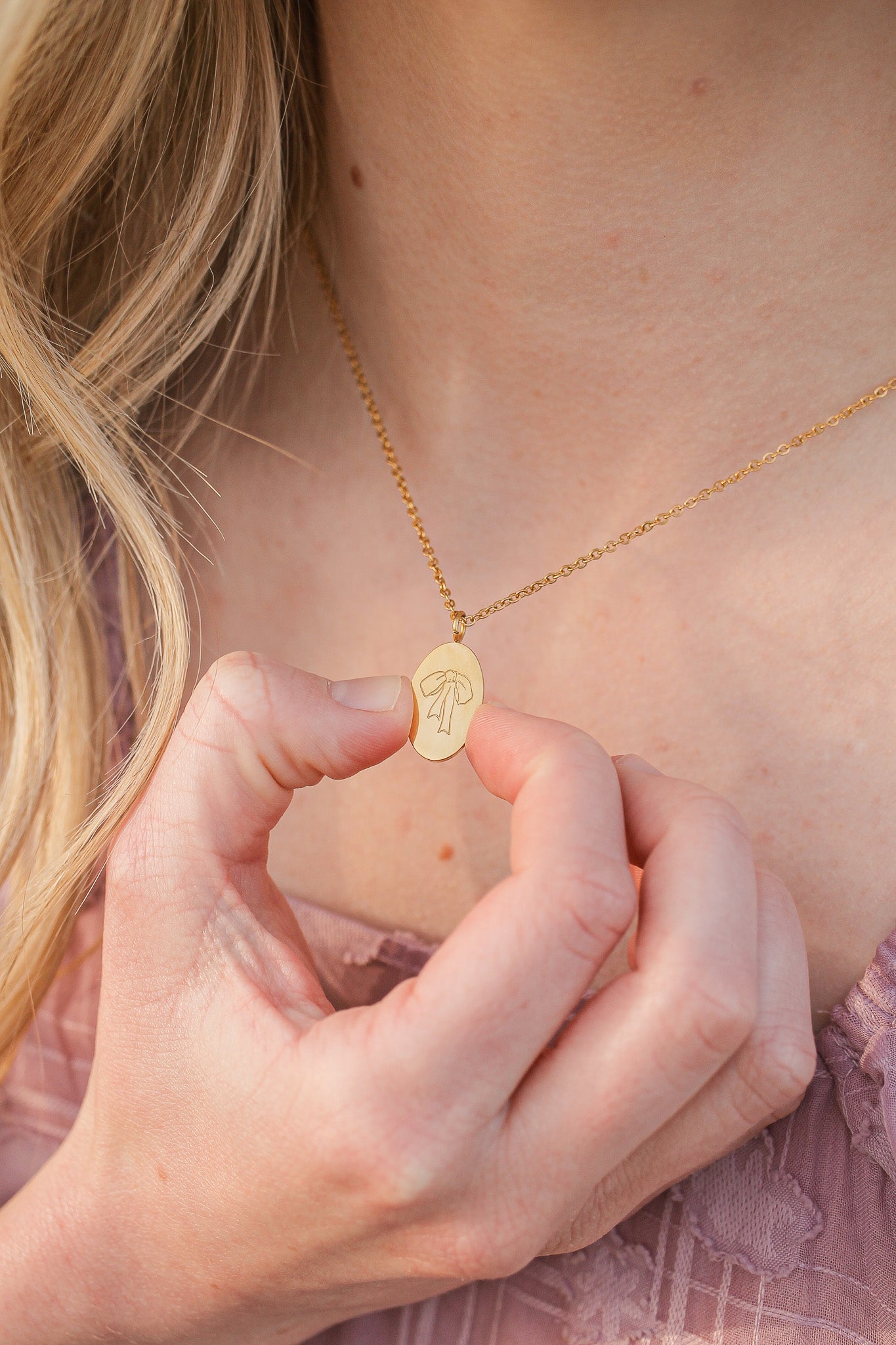 A person holds a gold necklace pendant featuring an engraved design, gazing at it attentively. The backdrop suggests a soft, natural light, emphasizing a serene atmosphere.