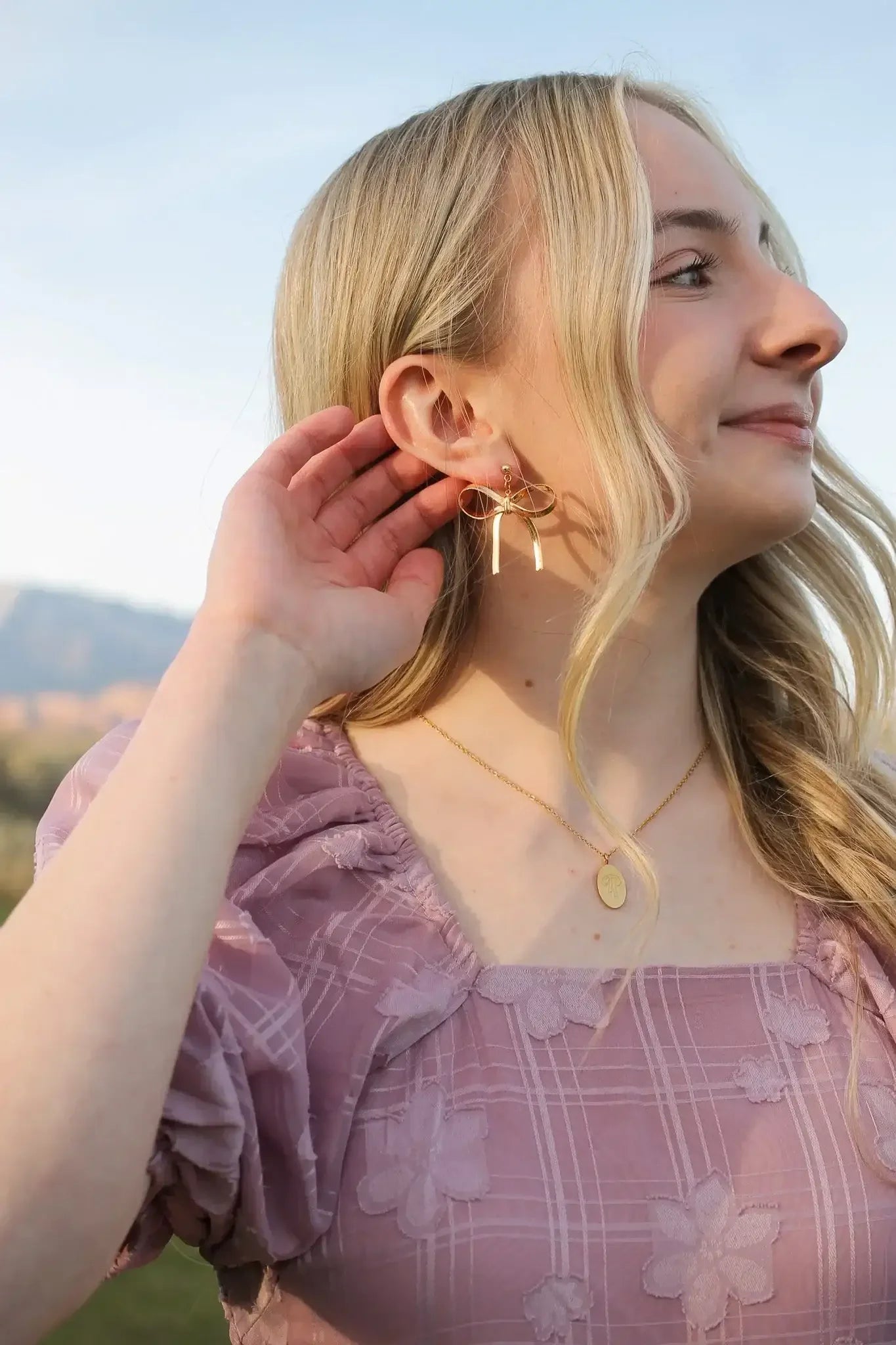 A woman with blonde hair touches her ear, showcasing a gold bow-shaped earring. She wears a pink blouse with floral patterns, set against a clear sky and distant mountains.