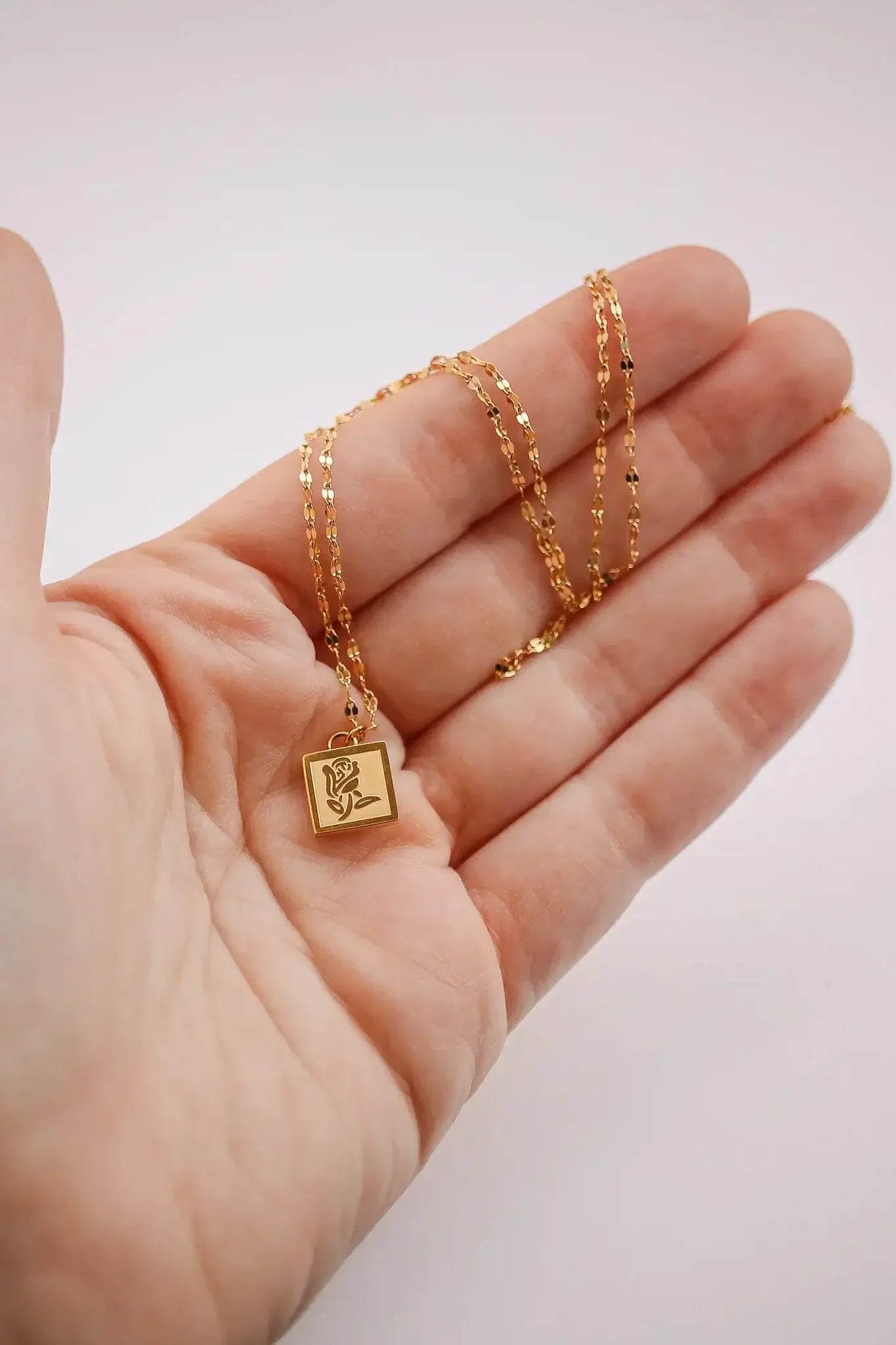 A hand holds a delicate gold necklace with a square pendant featuring a rose design, showcasing the jewelry against a light-colored background.